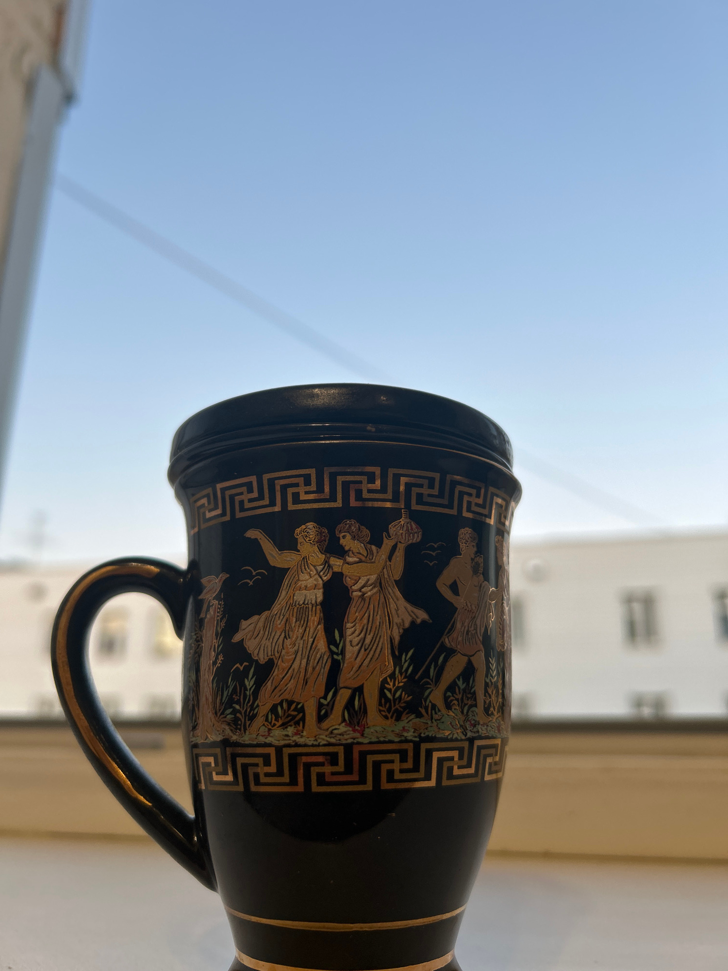 An ornate black mug with gold detailing sits on a windowsill, with a bright blue sky and a blurred cityscape visible in the background. The mug features classical figures in gold, reminiscent of ancient Greek art, and is adorned with a Greek key border. A window frame is visible on the left side of the image.