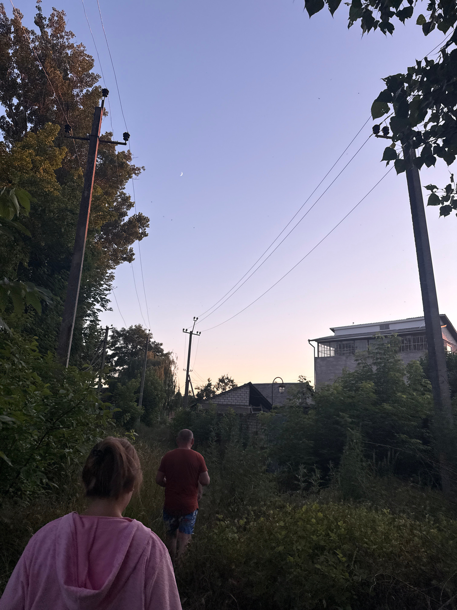 In Obukhivka, Ukraine, two people walk along a path lined with dense vegetation and trees, with utility poles and power lines visible. The sky is clear and fades from a pale blue at the top to a warm orange hue near the horizon. A crescent moon is faintly visible in the upper left portion of the sky. In the background, several buildings and structures can be seen peeking through the foliage. The person in the foreground is wearing a pink hooded sweatshirt, and the person in the background is wearing a red t-shirt and patterned shorts. They appear to be walking away from the camera into the overgrown path.