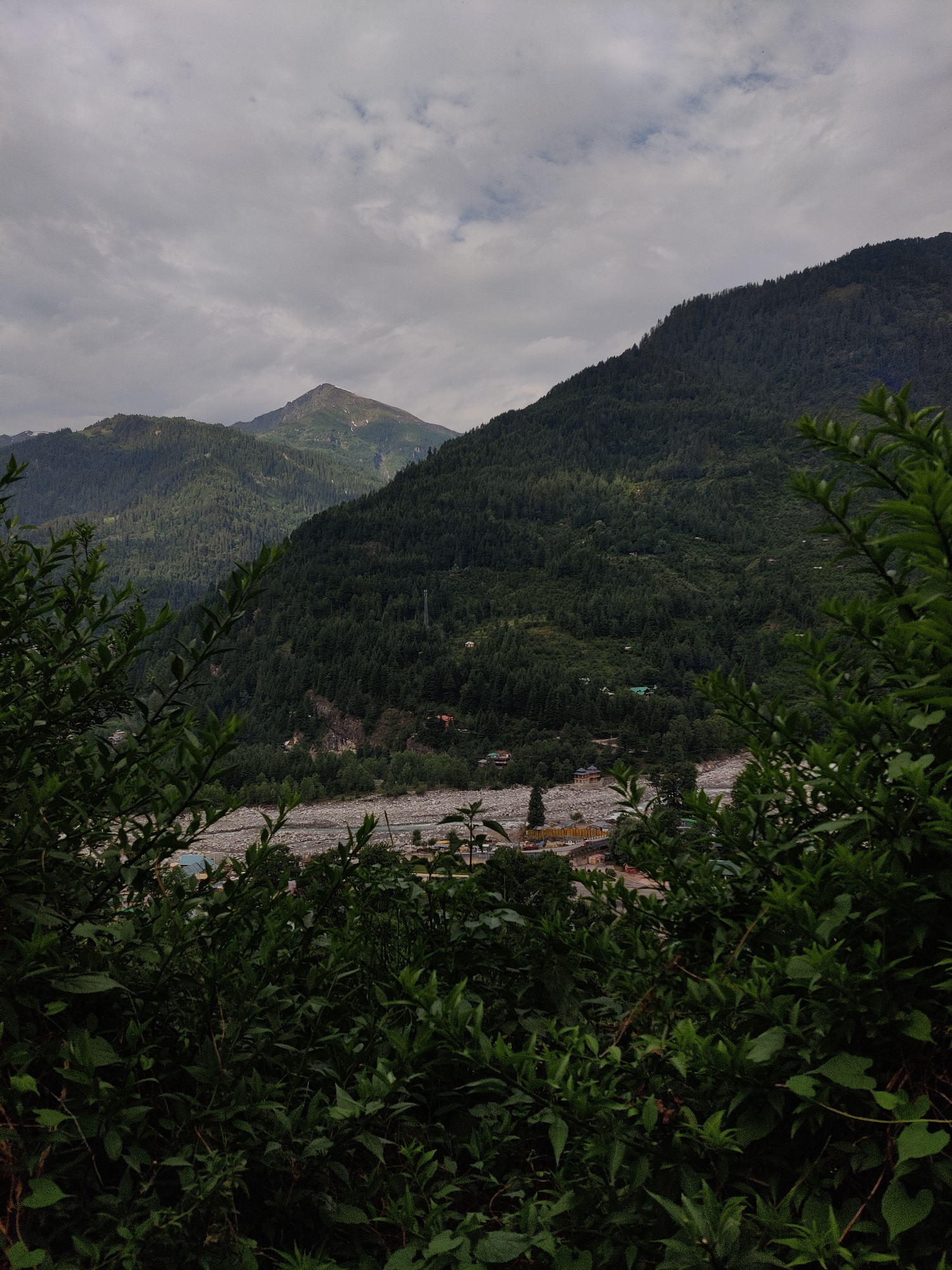 This image depicts a mountainous landscape viewed from a slightly elevated position, with dense green foliage in the foreground framing the scene. In the background, a riverbed filled with rocks runs through a valley, and beyond that, the slopes of mountains covered in coniferous trees rise towards a cloudy sky. A prominent, pointed peak, partially illuminated, stands out against the overcast horizon. Several small buildings, some with green roofs, are scattered across the lower slopes of the mountains, suggesting a small settlement or village. A telecommunications tower is visible on one of the slopes. The overall impression is one of a serene, natural environment with signs of human habitation integrated into the landscape. The location is identified as Vashisht, India, suggesting a Himalayan region known for its natural beauty and potentially spiritual significance, given Vashisht's association with hot springs and temples.