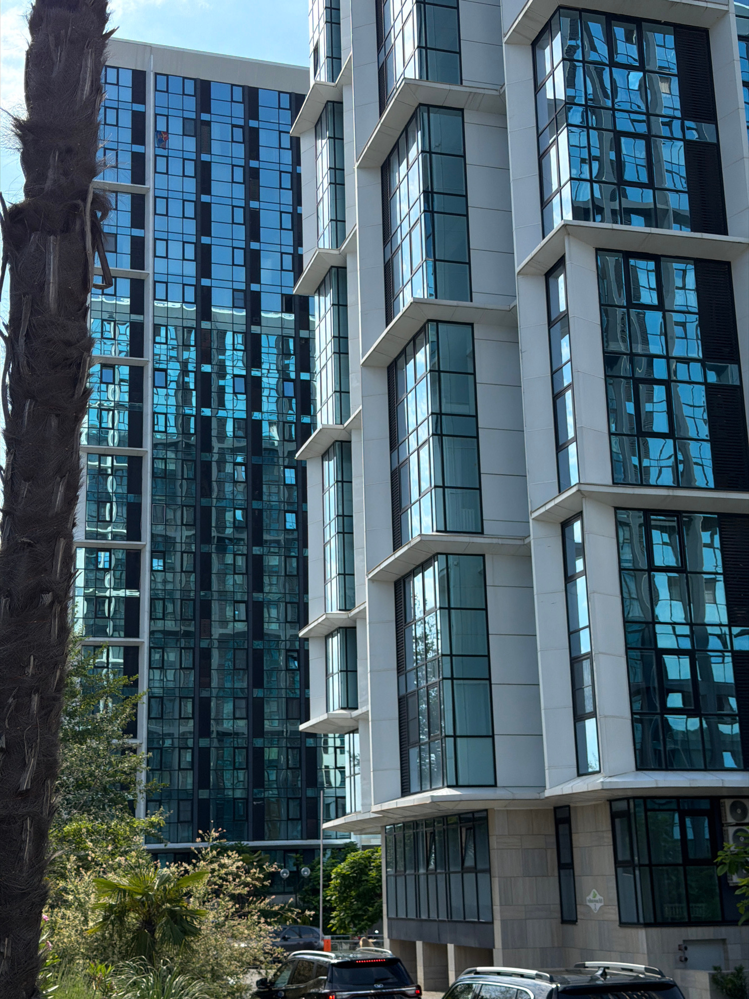 This image depicts a modern residential complex with a distinctive architectural style, characterized by prominent balconies and extensive use of glass facades. The buildings are tall and appear to be newly constructed, reflecting the surrounding environment and sky. The scene is set against a bright, slightly overcast sky, suggesting it is daytime.
In the foreground, a portion of a palm tree trunk is visible on the left, its rough texture contrasting with the smooth surfaces of the buildings. Further back, lush green trees and foliage create a natural boundary between the street and the residential structures. Several cars are parked along the street, indicating an urban or suburban setting. The architectural design of the buildings, with their clean lines and reflective windows, suggests a contemporary aesthetic often found in well-developed urban areas.
While the image does not explicitly show signage indicating "Sochi, Russia," the presence of palm trees and the overall climate suggested by the greenery and bright sky are consistent with the subtropical environment of Sochi. The modern architecture is also in line with the rapid development and modernization that has taken place in Sochi in recent years, particularly following its hosting of the 2014 Winter Olympics. The buildings' design, with cantilevered sections and large glass panels, is typical of contemporary residential developments aimed at maximizing natural light and offering scenic views, which would be desirable in a coastal city like Sochi.