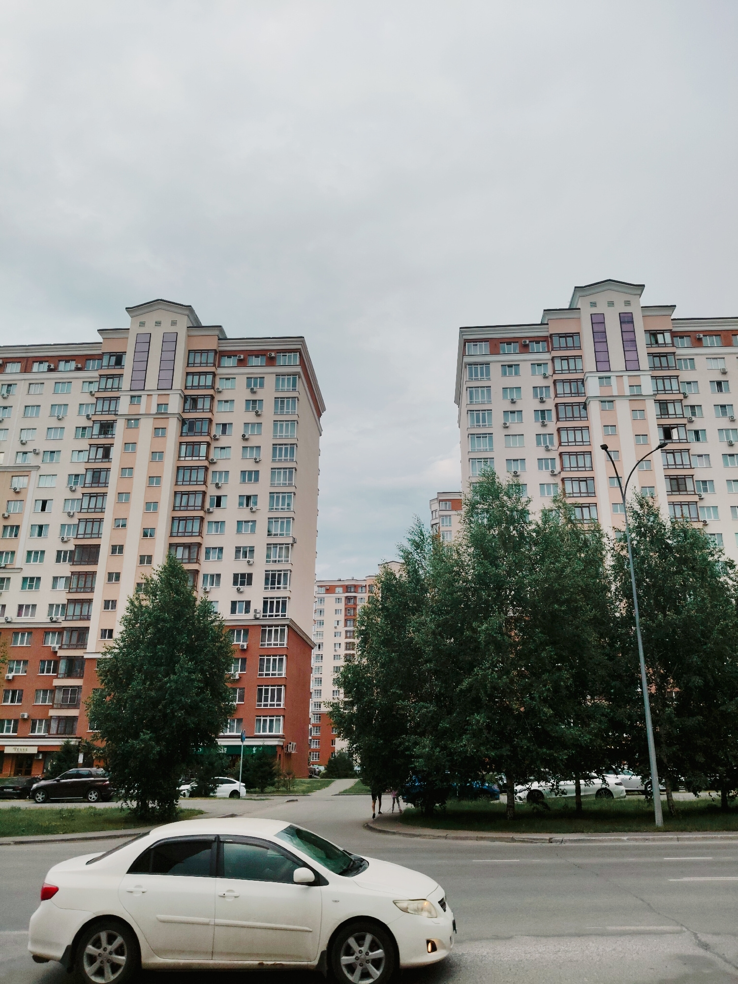 The image depicts a street view in Kemerovo, Russia, with two tall, modern apartment buildings dominating the background. The buildings are light-colored with cream and light pink facades, featuring numerous windows and balconies. The sky is overcast and gray. In the foreground, a white sedan is driving on a paved road. Trees with dense green foliage line the street between the road and the buildings, obscuring some of the lower portions of the apartment blocks. In the distance, another apartment building, similar in style, is visible. The overall scene suggests a residential area in a city.