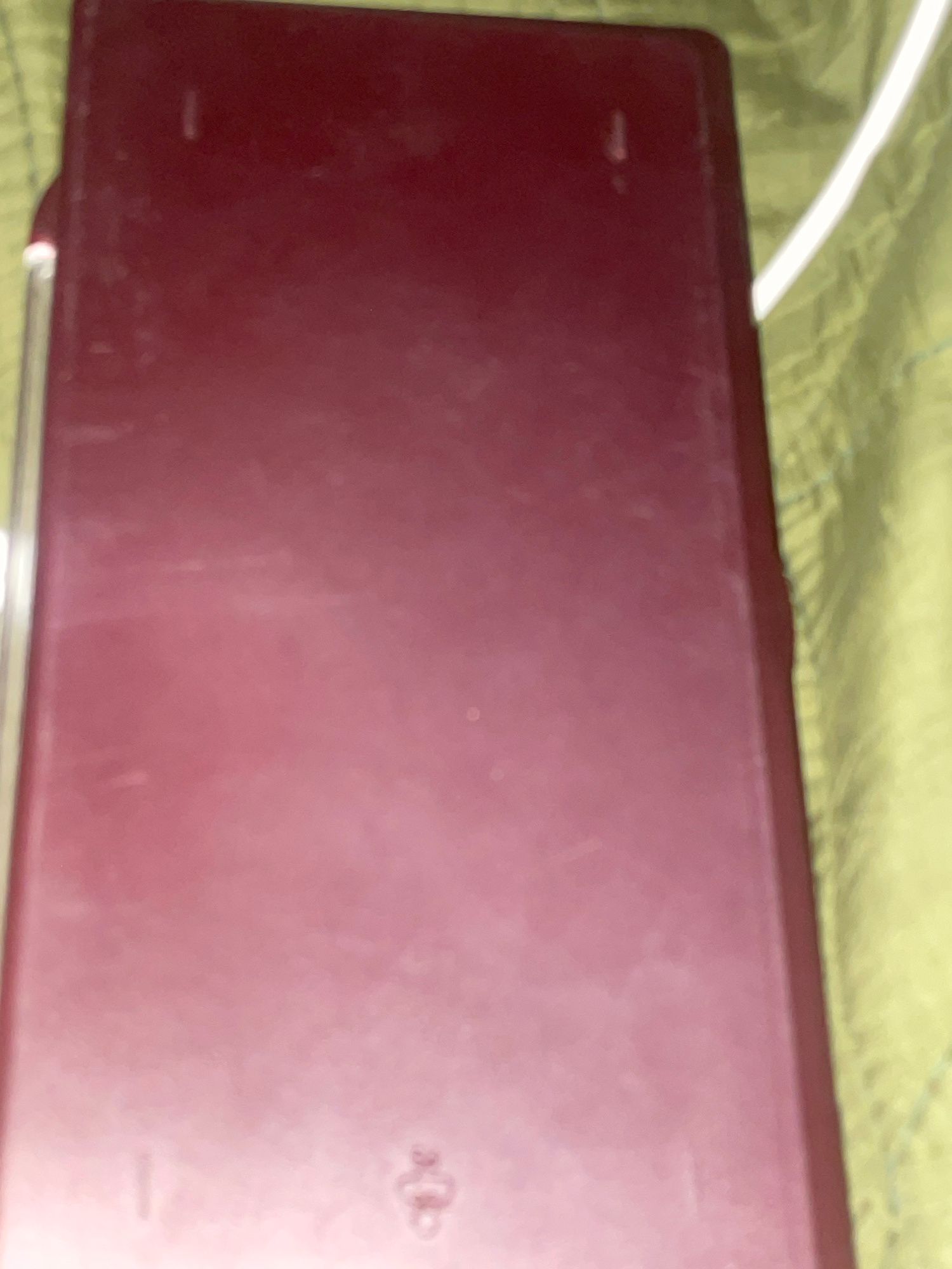 The image shows a close-up, angled view of a dark red, rectangular object, possibly a case or cover, resting on a light green, textured surface. The object appears to be made of plastic and has a matte finish, with some faint scuff marks visible. There is a thin, white cable running along the right edge of the frame, partially obscuring the object. The background suggests a casual, indoor setting. The image does not provide any information about the city of Jalingo, Nigeria, or any specific location within it.