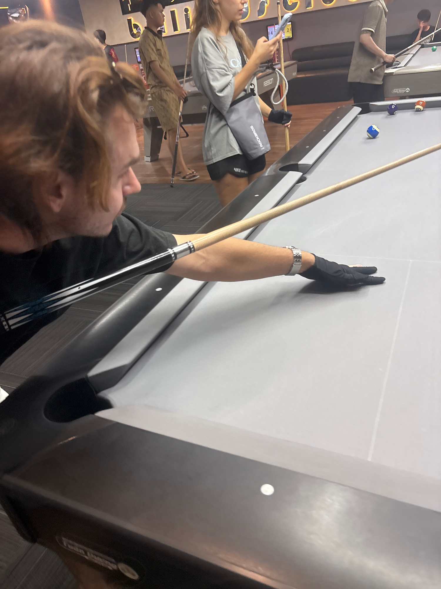 This image shows a group of people in a dimly lit establishment, possibly a bar or entertainment venue, with several pool tables. The scene is oriented vertically, so the people and objects appear sideways.
In the foreground, a person with shoulder-length blonde hair is in focus, wearing a black t-shirt and looking downwards. They are holding a pool cue. Another person, wearing a grey t-shirt and black shorts, is visible in the background, holding a pool cue and standing near a pool table. They are also wearing black gloves. A grey bag with the word "ROCKEROS" and "NIKE" is on the floor near them. Further in the background, another person in a patterned shirt is seen with a pool cue.
Several pool balls are scattered on a pool table in the midground, including a blue and white striped ball and a solid orange ball. The pool table is covered with a light grey felt. The setting appears to be indoors, with wooden flooring and some decorative lighting visible. The overall mood seems relaxed and casual, suggesting a social gathering or recreational activity. No specific time of day or weather conditions can be determined from the image. The location context provided is Đà Nẵng, Vietnam.