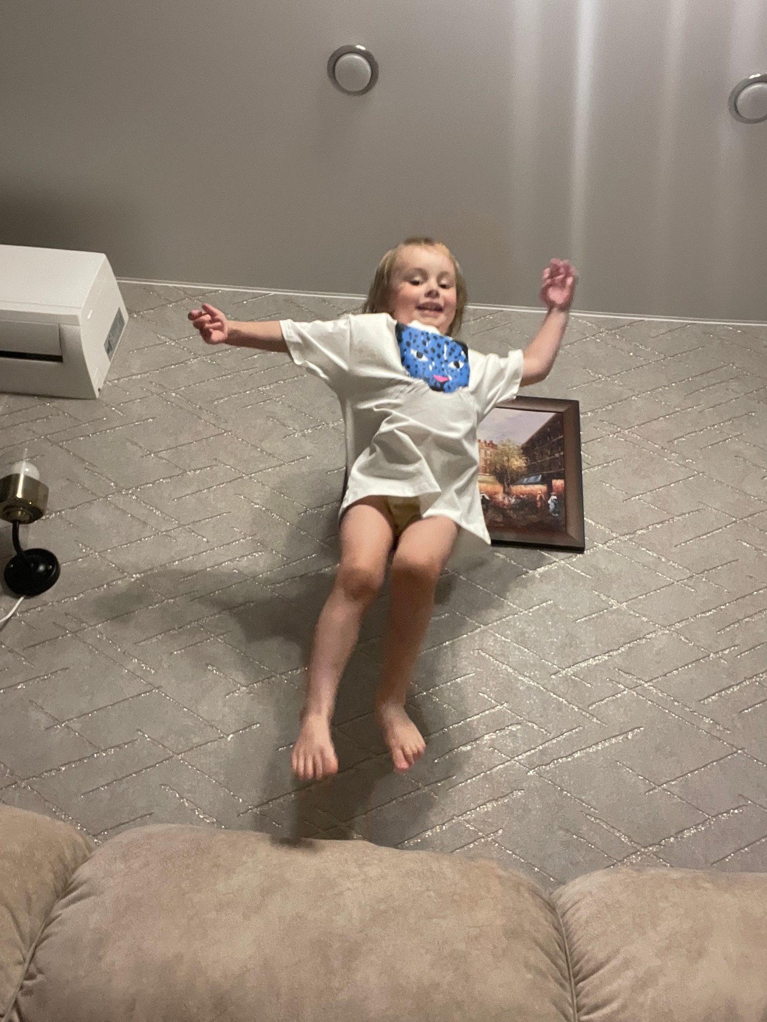 This media file features a young child in mid-air, seemingly jumping or falling, captured in a vertical orientation. The child, with blonde hair and wearing a white t-shirt with a blue bib featuring a patterned design, is positioned against a textured wallpaper. The wallpaper has a grey background with a repeating geometric pattern of thin, metallic lines.
To the left of the child, a circular light fixture is visible on a plain grey wall. Below the child, a white rectangular object, possibly an air purifier or a humidifier, sits on the floor next to a small lamp with a metallic base and a black cord. In the upper part of the image, a framed picture hangs on the wallpapered wall, depicting a scene with figures and trees. To the right, a portion of a large, soft beige sofa is visible, suggesting an indoor domestic setting, likely a living room.
The child's expression is somewhat obscured, but their arms and legs are spread wide, conveying a sense of movement and perhaps excitement or surprise. The lighting in the room appears to be artificial. No specific time of day or weather conditions can be determined from the image. The location context provided is Vladivostok, Russia, but there are no explicit visual cues within the image to confirm this. No visible text is present in the media file.