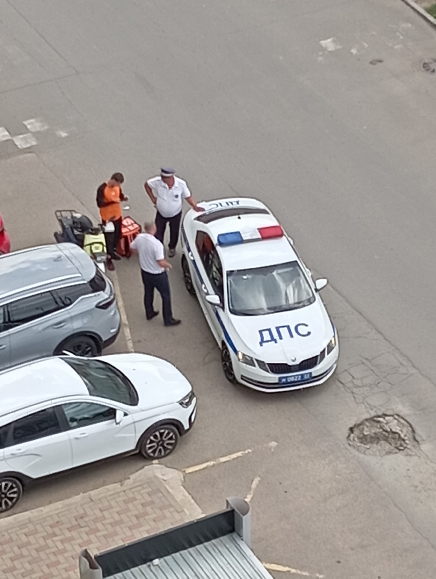 This aerial view captures a scene on an asphalt surface, possibly a parking lot or street, in Krasnodar, Russia. The time of day appears to be daytime, with overcast conditions.
The primary subjects are several people and vehicles. A white police car, marked with blue Cyrillic text "ДПС" on the hood and a license plate with "М 0822 М", is parked centrally. It has a red, white, and blue light bar on its roof.
Interacting with the police car are at least three individuals. One person in a white uniform and a blue cap appears to be speaking to another person dressed in an orange vest, who is lying on the ground. A third person, also in a white shirt and dark pants, is standing nearby. A motorcycle with a yellow vest and a medical bag is also present.
Beyond the police car, two other white cars are visible. One is parked to the right of the police car, and another is in the foreground. A section of a building with horizontal siding is visible on the right edge of the image. The ground shows some wear and tear, including cracks and patches of discolored asphalt. The overall impression is of a police-related incident, potentially an accident or a medical emergency.