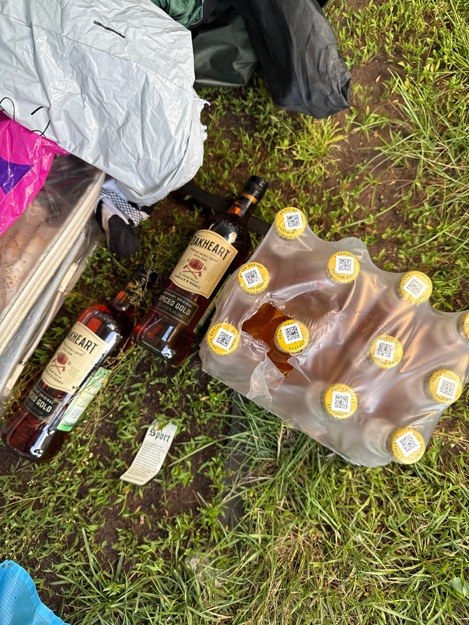 On the ground, which appears to be soil with sparse grass, are several items. Two bottles of "Oakheart Spiced Gold Premium Spirit Drink with Rum" are visible, lying on their sides. One bottle's label indicates 35% alc/vol. Next to these is a package containing multiple bottles of beer, enclosed in clear plastic packaging with yellow caps featuring QR codes and Cyrillic text. A white tag with the word "Sport" printed on it is also present on the ground. In the background, there are large, crumpled white and black plastic bags, and a green fabric item. The scene appears to be outdoors.