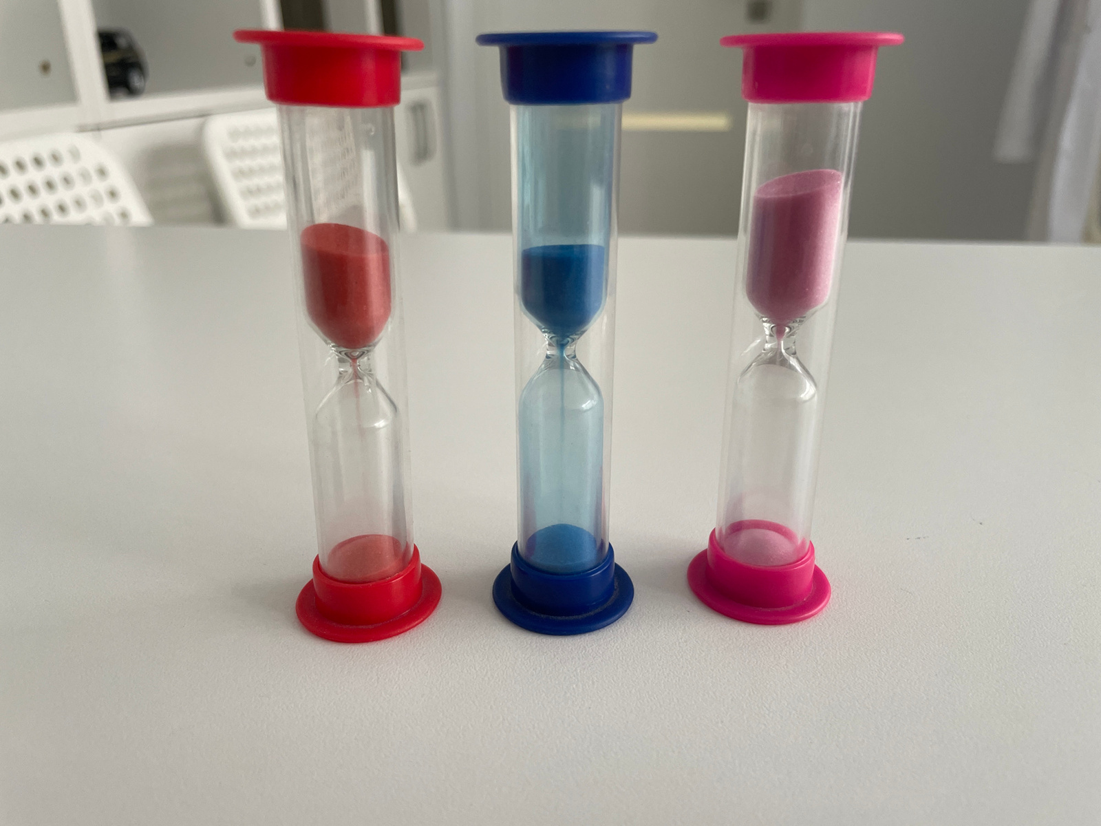 The image shows three hourglasses standing on a white surface, possibly a table. The hourglasses are of different colors and contain colored sand. From left to right, the first hourglass is red, filled with red sand. The second hourglass is blue, containing blue sand. The third hourglass is pink, filled with pink sand. The sand in the red and pink hourglasses is mostly in the top bulb, indicating that time has not yet begun to run or has just started. The blue hourglass shows some sand in the bottom bulb, suggesting that some time has already passed. The background is blurred, but suggests an indoor setting with some furniture, possibly chairs, visible. The lighting suggests it is daytime. There is no visible text in the image. The overall scene is simple and focuses on the hourglasses as the main subjects.