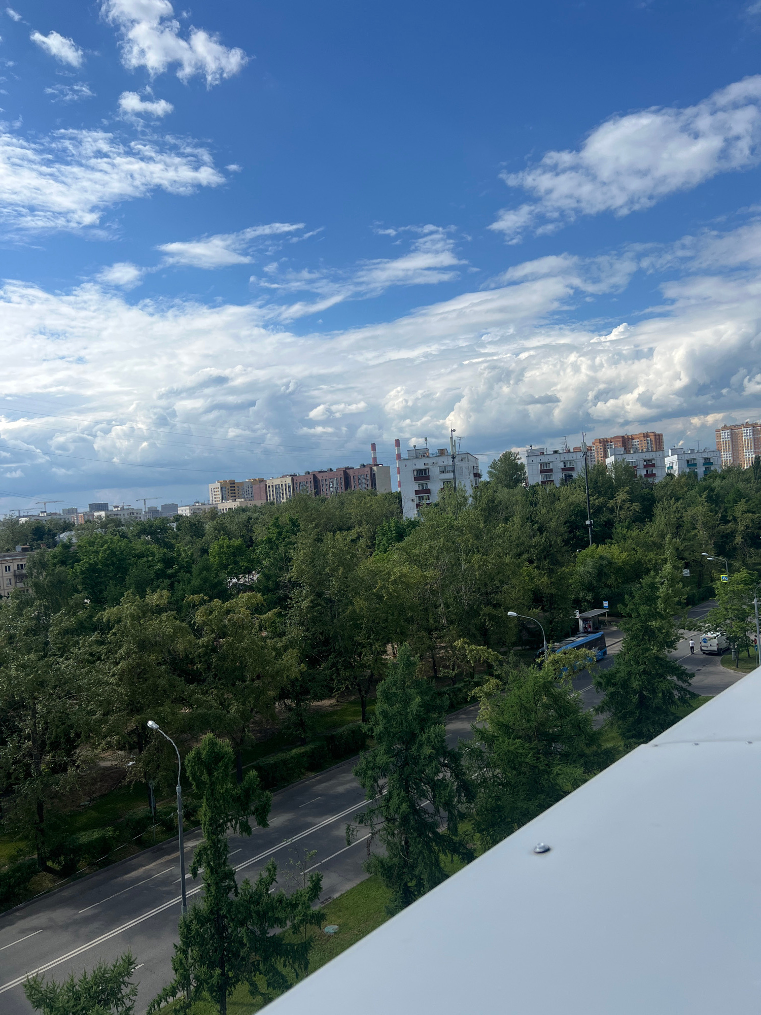 The image captures a view from an elevated position, looking down at a city street lined with trees and buildings. The scene is oriented vertically, suggesting the photo was taken with the camera rotated. The sky is a vibrant blue with scattered white clouds, indicating daytime and fair weather.
The foreground is dominated by a white, angled structure, likely a building's facade or a balcony railing, with visible fasteners. Below and extending into the background is a dense row of lush green trees, forming a canopy over the street. The street itself is paved and appears to be a two-lane road with white markings. There are lampposts along the street, one of which is illuminated, suggesting it might be late afternoon or early evening, or perhaps a daytime light.
Visible in the middle ground and background are residential buildings of varying heights and architectural styles. Some are modern, with multiple stories and balconies, while others have a more industrial appearance with visible pipes and ventilation shafts. A blue bus is stopped at a bus stop along the street, and a white vehicle is seen further down the road. A few individuals can be seen on the sidewalk or near the bus stop, but they are not a primary focus.
The overall impression is of a typical urban environment in Moscow, Russia, likely during the warmer months given the greenery. The composition highlights the contrast between the natural elements of trees and sky and the man-made structures of buildings and infrastructure. There is no visible text in the image.
