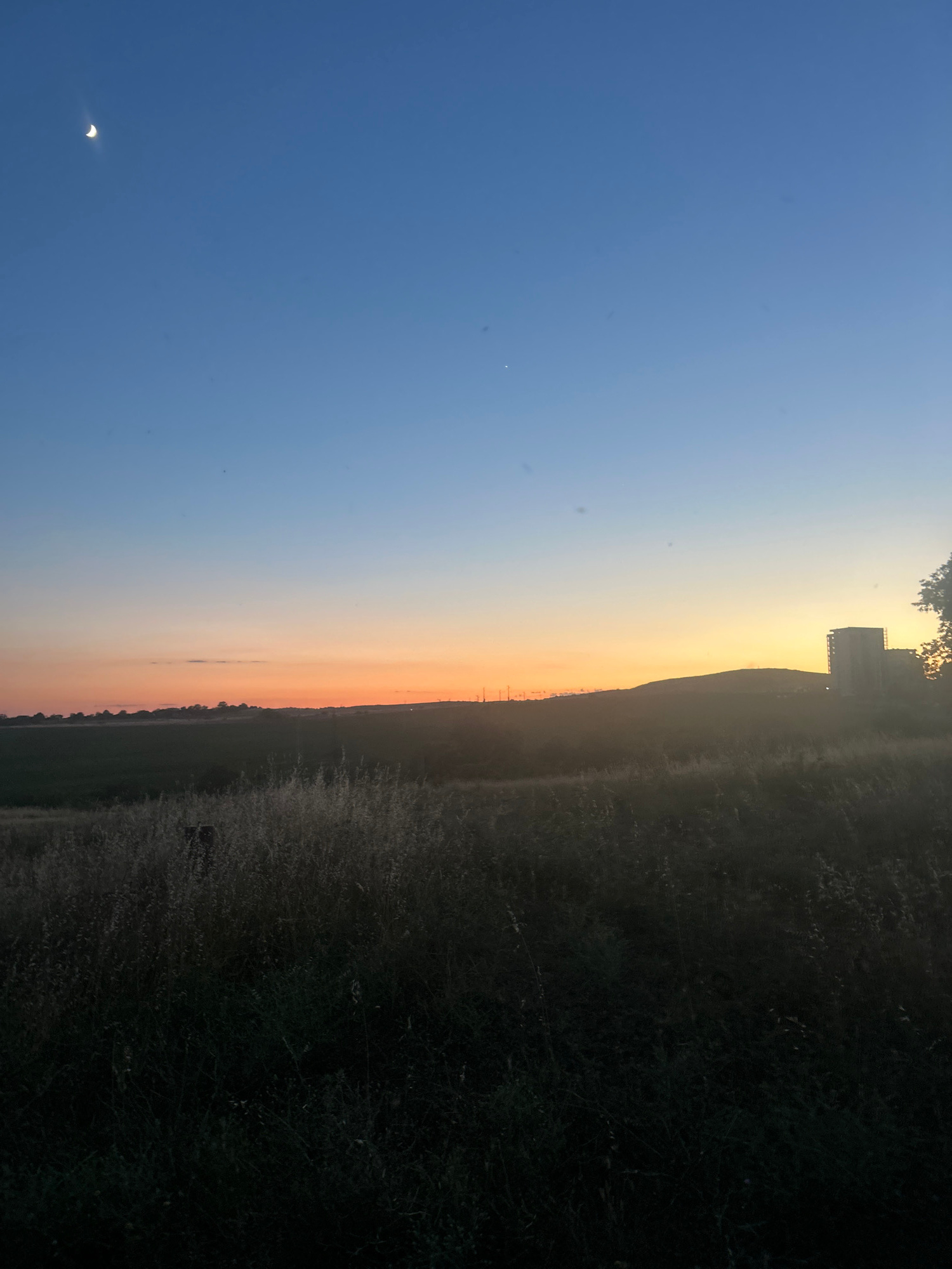 A twilight scene captures a wide, open field under a gradient sky transitioning from deep blue at the top to warm orange and pink hues near the horizon. A crescent moon is visible in the upper left quadrant of the sky. A faint star is also present in the upper right quadrant. In the middle ground, a gently sloping hill is silhouetted against the vibrant sunset colors. On the crest of the hill, a modern, multi-story building, possibly under construction or with scaffolding, stands out. The foreground is dominated by a dark, dense field of tall, dry grass, with a dark, indistinct object, possibly a post or a fence, partially obscured within it. The overall impression is a rural or semi-rural landscape, likely on the outskirts of a city.