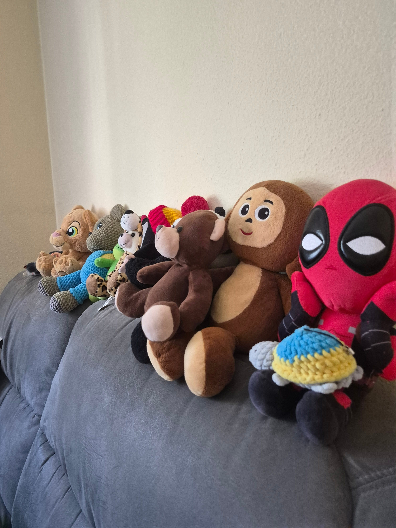 An overhead, eye-level, medium shot captures a collection of stuffed animals lined up on a grey couch against a light-colored wall. The stuffed animals, from left to right, include a lion cub, a blue-knitted lion, a green-knitted creature, a spotted giraffe, a black and white cat, a brown bear, a monkey, a Deadpool character, and a small turtle with a blue and yellow crocheted shell. The Deadpool character is positioned to the right of the monkey and is slightly turned towards the viewer. The turtle is placed in front of the Deadpool character. The couch is upholstered in a textured grey fabric, and its cushions are visible. The wall behind the couch is a plain, light color.