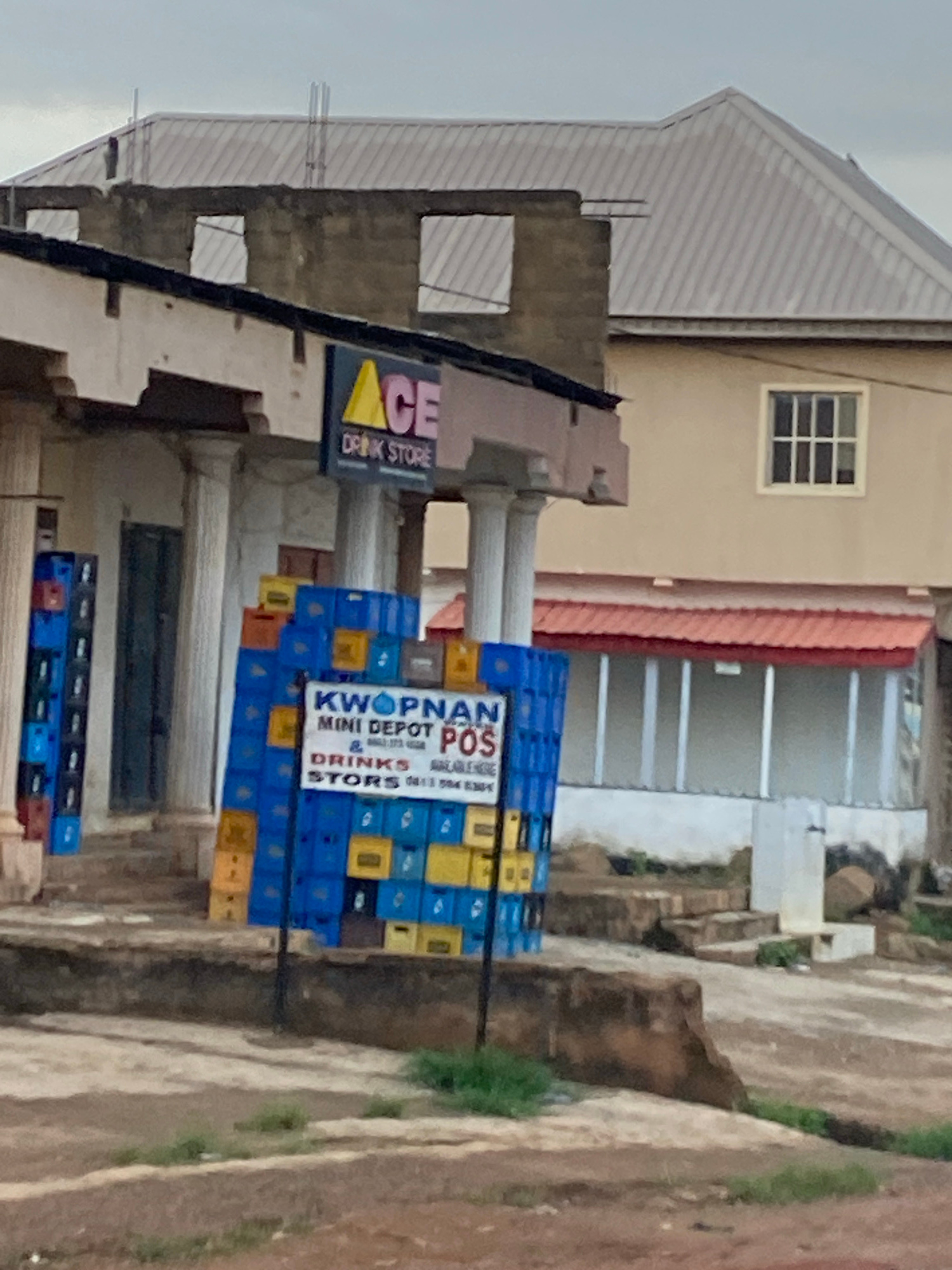 An outdoor, slightly elevated view shows a building under construction next to a completed building with a red awning. In the foreground, a stack of colorful crates, primarily blue and yellow, is visible. A sign with the text "KWOPNAN MINI DEPOT POS & DRINKS STORS" is attached to the crates. To the left, more crates are stacked vertically against a wall. A sign above a doorway on the left building reads "ACE DRINK STORE". The ground in front of the buildings is unpaved and appears worn. The overall impression is one of a commercial area in a state of development. The sky is overcast. The scene is consistent with a street view in Jos, Nigeria.