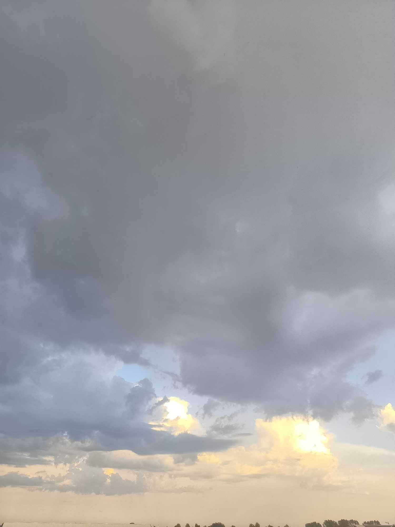 A low-angle, full-frame shot captures a dramatic, cloudy sky over Uithoorn, Netherlands. The clouds are predominantly dark gray, with lighter patches and streaks indicating varying thickness and light penetration. In the lower half of the image, shafts of bright, yellowish-white light break through the clouds, suggesting the sun is low on the horizon or partially obscured. A faint, light-colored horizon line, possibly indicating water or land, is visible at the very bottom of the frame. Silhouetted against the sky at the bottom are indistinct, dark shapes that could be trees or buildings, characteristic of the Uithoorn skyline. No people or specific objects are clearly discernible in the sky. The overall impression is one of an approaching storm or the aftermath of one, with a dynamic interplay of light and shadow.