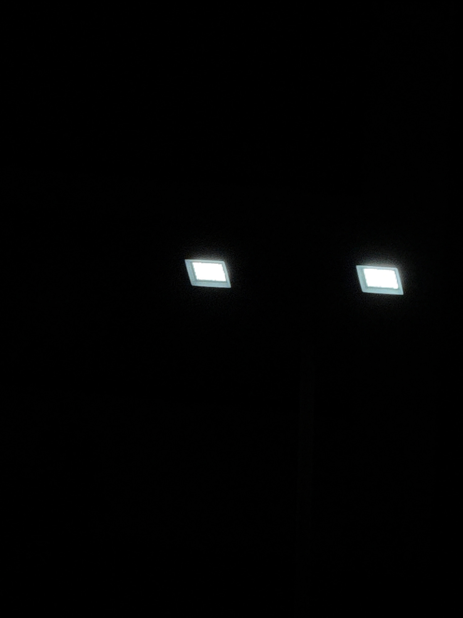 The image displays a dark, abstract scene with two bright, rectangular lights positioned on the right side. The lights appear to be illuminated from within, casting a soft glow on the surrounding darkness. One light is positioned slightly higher and further to the right than the other. The context of Tbilisi, Georgia is not discernible from the provided image.