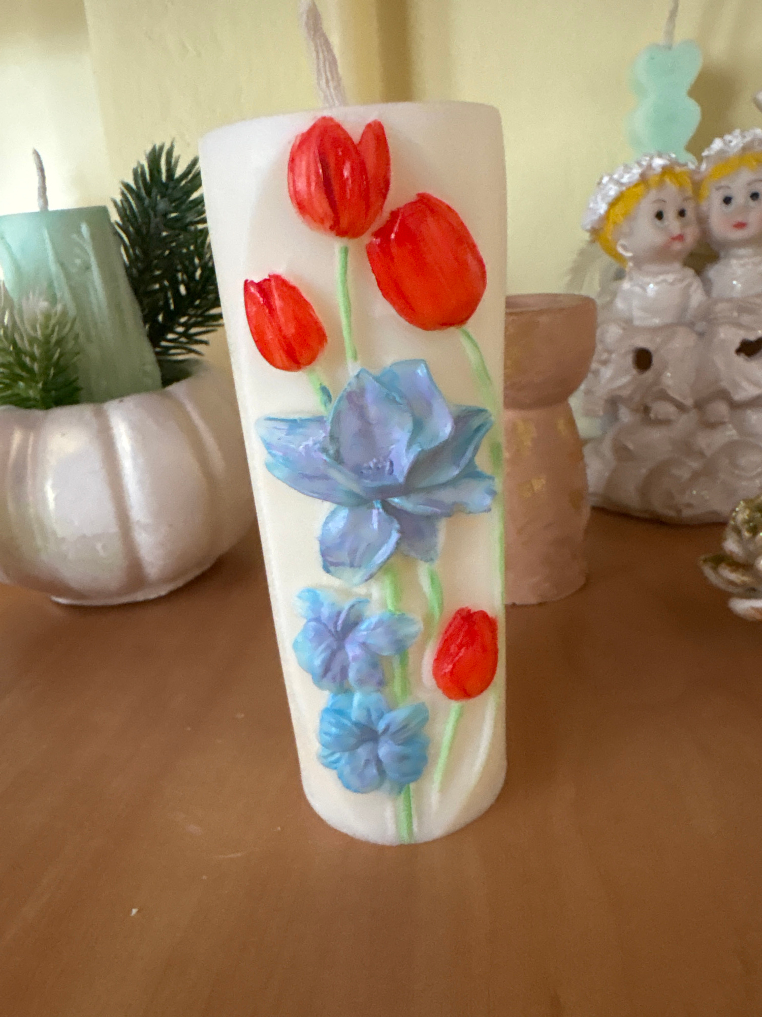 A vertically oriented, medium close-up shot features a white cylindrical candle with a floral design. The candle is adorned with red tulips and blue flowers, along with green stems. To the left of the candle, a pale green, Christmas tree-shaped candle is visible, partially obscured by a small, white, pumpkin-shaped pot containing a miniature evergreen plant. In the background, two figurines of children in white attire with silver headbands are present, along with a light brown cylindrical object. The scene is set on a flat, brown surface, likely a table. The background is a solid, pale yellow wall. The image is taken in Ветрынскі сельскі Савет, Belarus.
