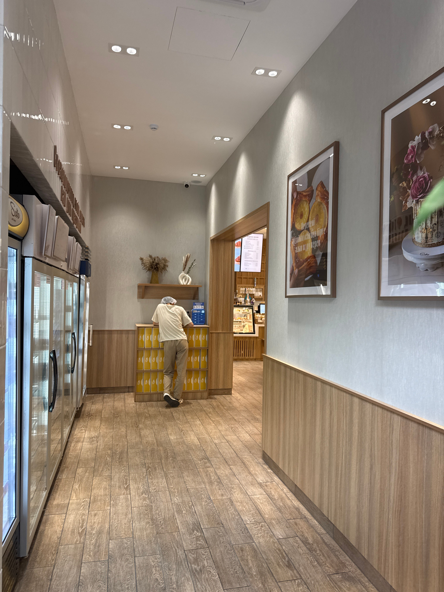 In a brightly lit bakery interior, a person dressed in casual clothing, including a light-colored long-sleeved shirt and khaki pants, is standing and interacting with a display of yellow boxes. The person is partially obscured by the boxes but appears to be reaching or examining them.
The setting is modern and clean, with light-colored walls and wooden flooring. Above, two framed pictures hang on the wall, one featuring a floral arrangement and the other displaying baked goods. The bakery counter is visible, with a menu board and refrigerated display cases. The overall atmosphere is calm and appears to be during business hours. The location is identified as Almaty, Kazakhstan.