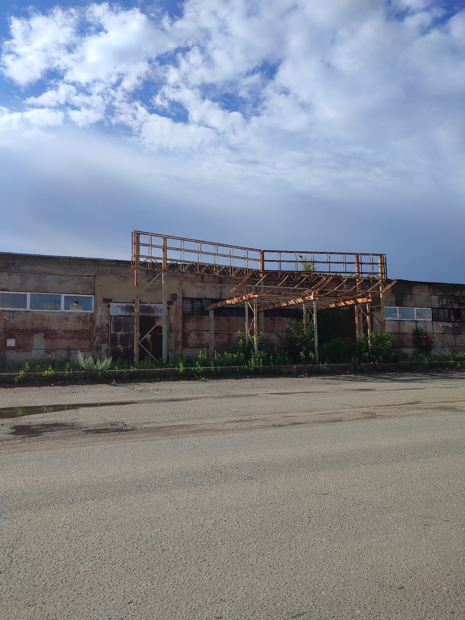 The image depicts a sunlit scene featuring an abandoned industrial building with a large, rusted metal structure extending from its facade. The building itself appears weathered and shows signs of disrepair, with patches of rust and peeling paint. Multiple windows, some with intact glass and others broken, are visible along its length. A large, corrugated metal door is also present, partially open to reveal a dark interior. Green vegetation is growing in front of the building, encroaching on the pavement. In the foreground, a paved road stretches across the width of the image, with several puddles reflecting the sky. The sky above is a mix of blue with scattered white clouds, some of which appear heavier and darker, suggesting potential for rain. The overall impression is one of urban decay and neglect in an industrial area. The location is identified as Orenburg, Russia.