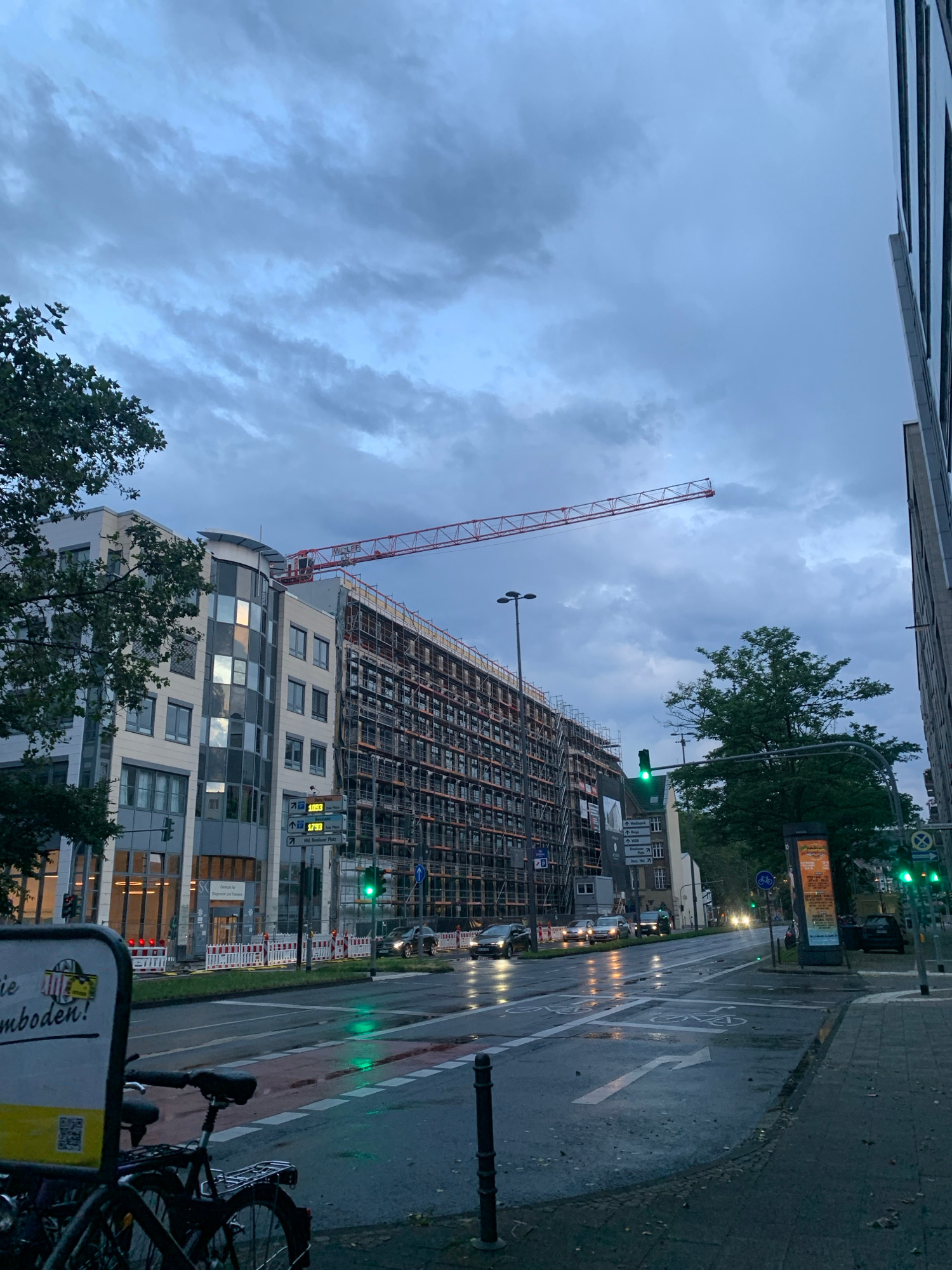 This image depicts a street scene at dusk, with a large building under construction dominating the view. A red construction crane extends from the building, reaching towards the overcast sky. The building itself is clad in a mix of modern materials, with large windows reflecting the dim light. Scaffolding covers much of the upper floors, indicating ongoing work.
In the foreground, a wet asphalt road curves, marked with white lines and bicycle symbols, suggesting a dedicated bike lane. The wetness indicates recent rain, and puddles reflect the streetlights and traffic signals. A bicycle is parked on the right side of the frame, partially obscuring a sign with some text on it. Traffic lights are visible, with one showing a green signal, implying that vehicles are allowed to proceed. A billboard with an advertisement is also present, and various street signs are scattered throughout the scene.
The overall atmosphere is muted, likely due to the low light conditions of dusk and the cloudy sky. The presence of construction suggests urban development, and the wet street indicates a recent or ongoing weather event. No people are clearly visible in the frame. The text on the sign in the foreground appears to be German.