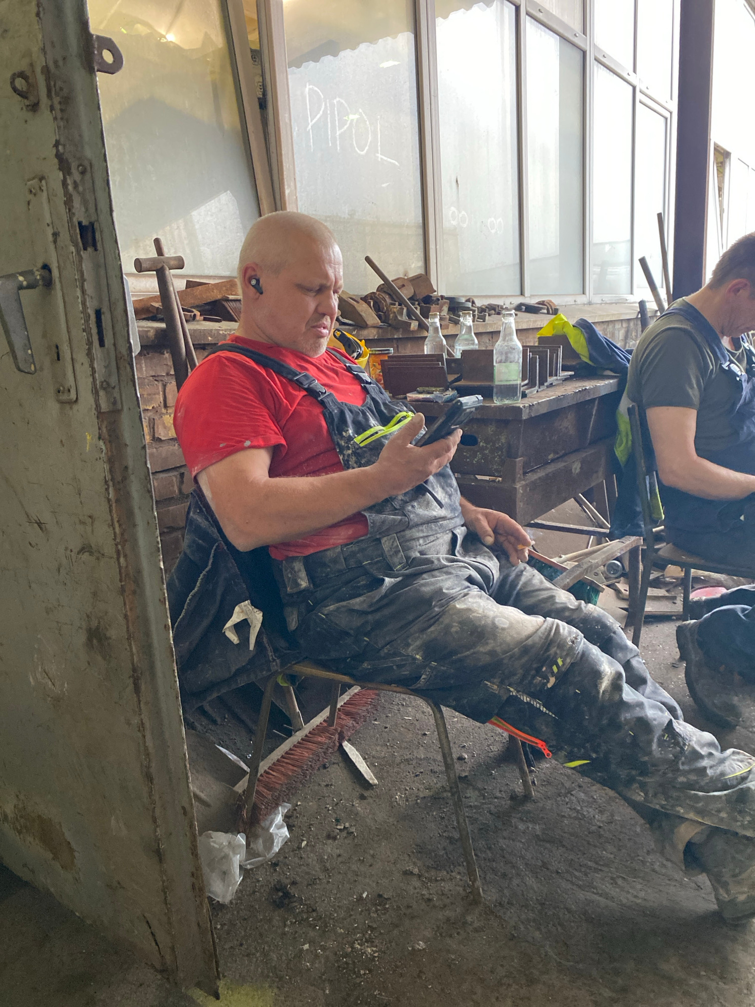 This image depicts two men in what appears to be a workshop or industrial setting. One man, bald and wearing a red t-shirt and overalls, is seated and looking at a smartphone. He has a hearing aid in his right ear. The other man is in the background, also wearing work clothes, and appears to be seated or leaning.
The scene is cluttered with various objects, including bottles, tools, a broom, and what looks like discarded materials. There are large windows with a grid pattern in the background, suggesting a well-lit indoor space, possibly a factory or a large workshop. The floor is concrete and dusty, consistent with an industrial environment.
The overall impression is of a work break or downtime. There is no clear indication of the time of day or weather conditions. The atmosphere seems casual, with the man on the phone engaged with his device.
Visible text includes "PPOL" written in white on a windowpane in the background. There are also some indistinct markings on the overalls of the seated man, likely paint or dirt. The setting, given the "Poland" location context, is likely a workplace in Poland.