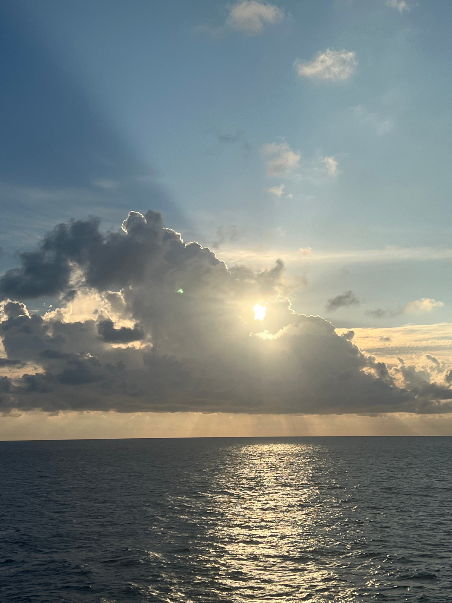 A dramatic sky over the open ocean features the sun peeking through a large, dark cloud formation. The sun's rays break through, creating a luminous glow that casts a shimmering reflection on the rippling water below. The ocean appears dark and vast, meeting the horizon under a gradient of blue sky and scattered clouds, some edged with light from the obscured sun.