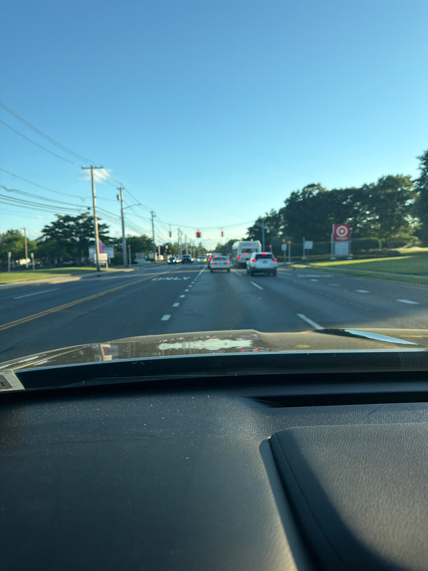The image shows a view from inside a vehicle, looking out through the windshield at a moderately busy road in Riverhead, New York. The sky is clear and blue. The road has multiple lanes, with cars driving in the same direction as the camera. Traffic lights are visible in the distance, and one is red, causing some of the cars to stop. On the right side of the road, there is a Target store sign, indicating the presence of a retail area. Trees line the sides of the road and the median, and utility poles with power lines are present on the left. The dashboard of the vehicle is visible in the foreground, with some dust particles on its surface.