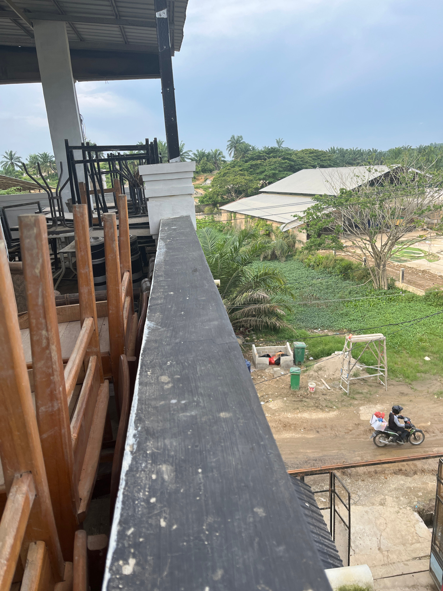 The image is a high-angle shot from a balcony or elevated structure looking out over a semi-urban or rural landscape, possibly in Benuaraja, Indonesia. In the foreground, stacked wooden chairs and tables are visible, suggesting an outdoor seating area of a restaurant or cafe. A dark, weathered railing runs along the edge of the foreground structure.
Beyond the immediate foreground, the scene opens up to a green, vegetated area with some buildings. A large, light-colored roofed structure, possibly a warehouse or factory, is prominent. Dense foliage, including palm trees, is scattered throughout the landscape.
In the middle ground, there's a construction or work area. A scaffolding structure stands near a pile of dirt and sand. Two green trash bins are present, and a person is seen lying down within a small concrete structure, possibly a makeshift shelter or a resting spot. Another person is riding a motorcycle with a box containing items on the back, moving along a dirt path.
The sky is overcast, indicating a cloudy or hazy day. The overall impression is of an active, yet somewhat unkempt, environment with elements of commerce, industry, and daily life.