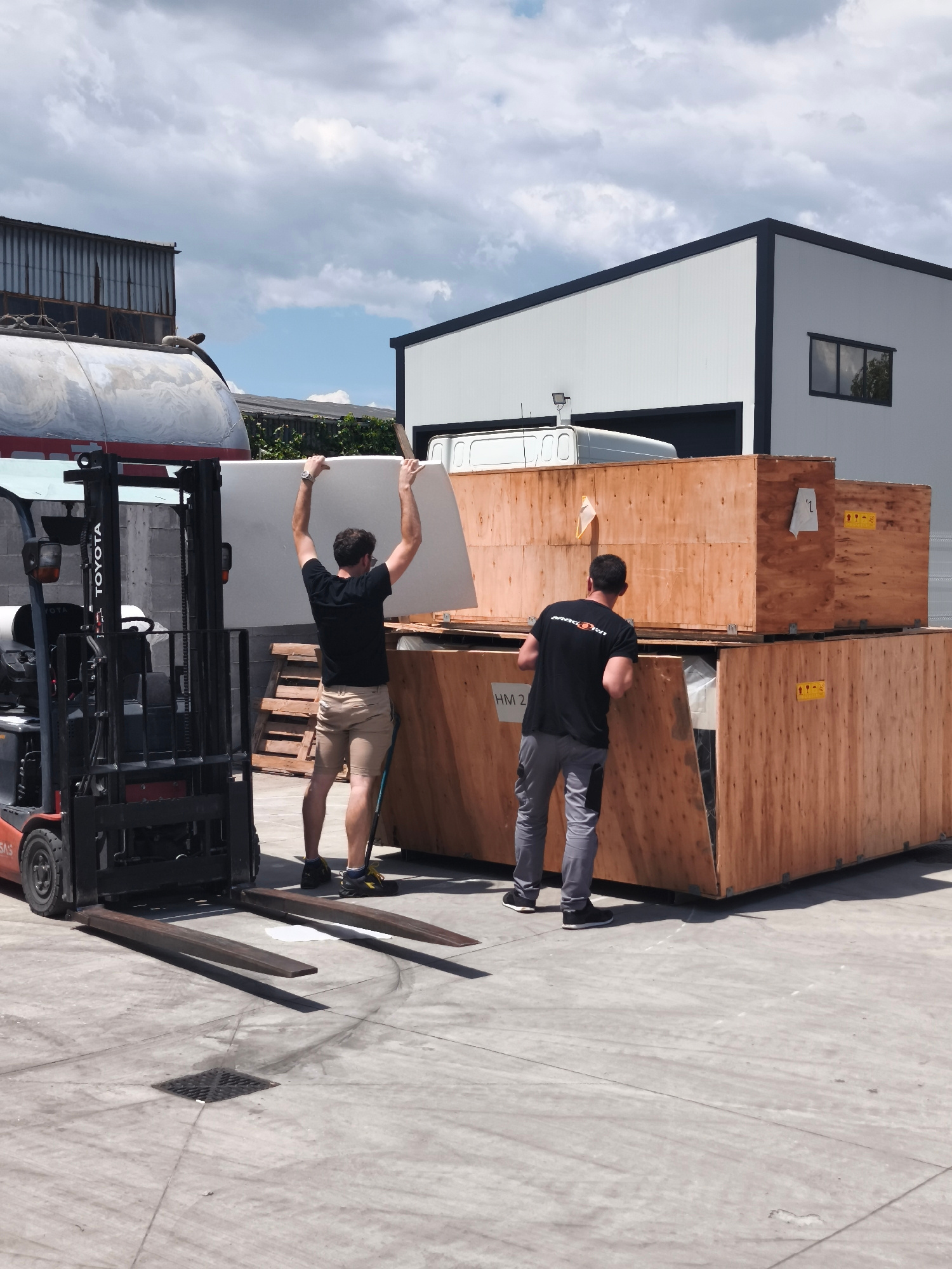 Two men are unloading a large white panel from a Toyota forklift. One man, wearing a black t-shirt and tan shorts, holds the panel above his head, while the other man, in a black t-shirt and gray pants, looks on from beside a stack of large wooden crates. The forklift is positioned on a concrete surface, and the scene appears to be an outdoor industrial or storage area with buildings in the background under a cloudy sky.