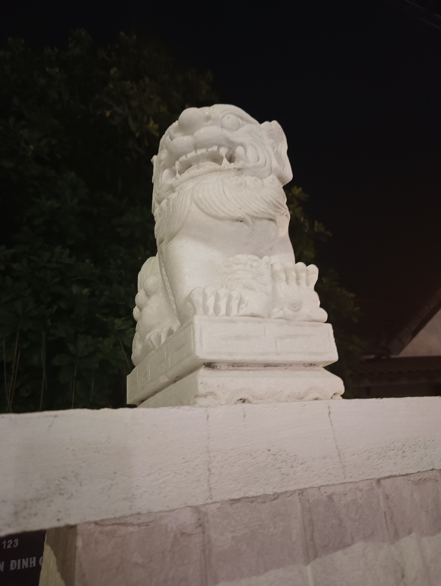 A white stone statue of a lion, likely a guardian figure, sits atop a white stone pedestal. The statue faces forward with its mouth open, revealing sharp teeth and a fierce expression. Its mane is depicted in carved waves, and its paws rest on the pedestal. The background consists of dark foliage, suggesting the image was taken at night, with some faint lighting illuminating the statue. The statue is positioned on a low white wall, with a portion of a building visible to the right. Below the wall, a sign with text "123 N DINH C" is partially visible, indicating a location in Phu Thuy Ward, Vietnam.