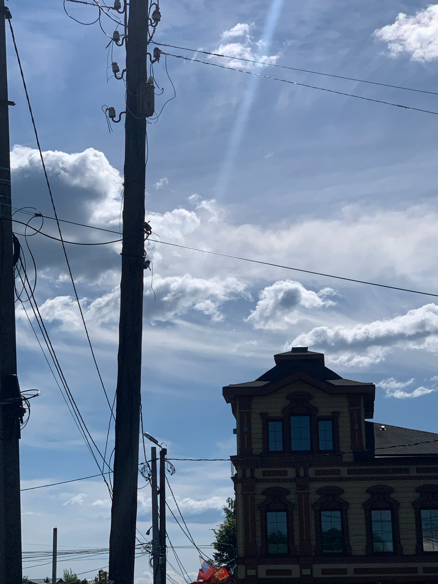 This is a vertically oriented, outdoor photograph taken from a low angle looking up at the sky. The dominant subjects are utility poles with numerous wires stretching across the frame, and a portion of an ornate, light-colored building with dark trim and several windows. The sky is a bright blue with scattered, wispy white clouds, suggesting a partly cloudy day. The sunlight creates a slight glare in the upper left quadrant of the image.
The utility poles are dark and appear to be made of wood, festooned with various insulators and electrical components. The building has detailed architectural features, including decorative window surrounds and a darker, possibly tiled roofline visible at the top. The perspective suggests the photo was taken in an urban or semi-urban environment where overhead wiring is prevalent.
The orientation of the image is rotated, making the building appear upside down relative to a typical upright orientation. There is no clear indication of people or any specific event taking place. The overall mood is one of an ordinary day, with the focus on the interplay between human infrastructure and the natural sky. The time of day appears to be daytime, likely morning or afternoon, given the brightness of the sky and the clarity of the clouds. No text is visible in the image.