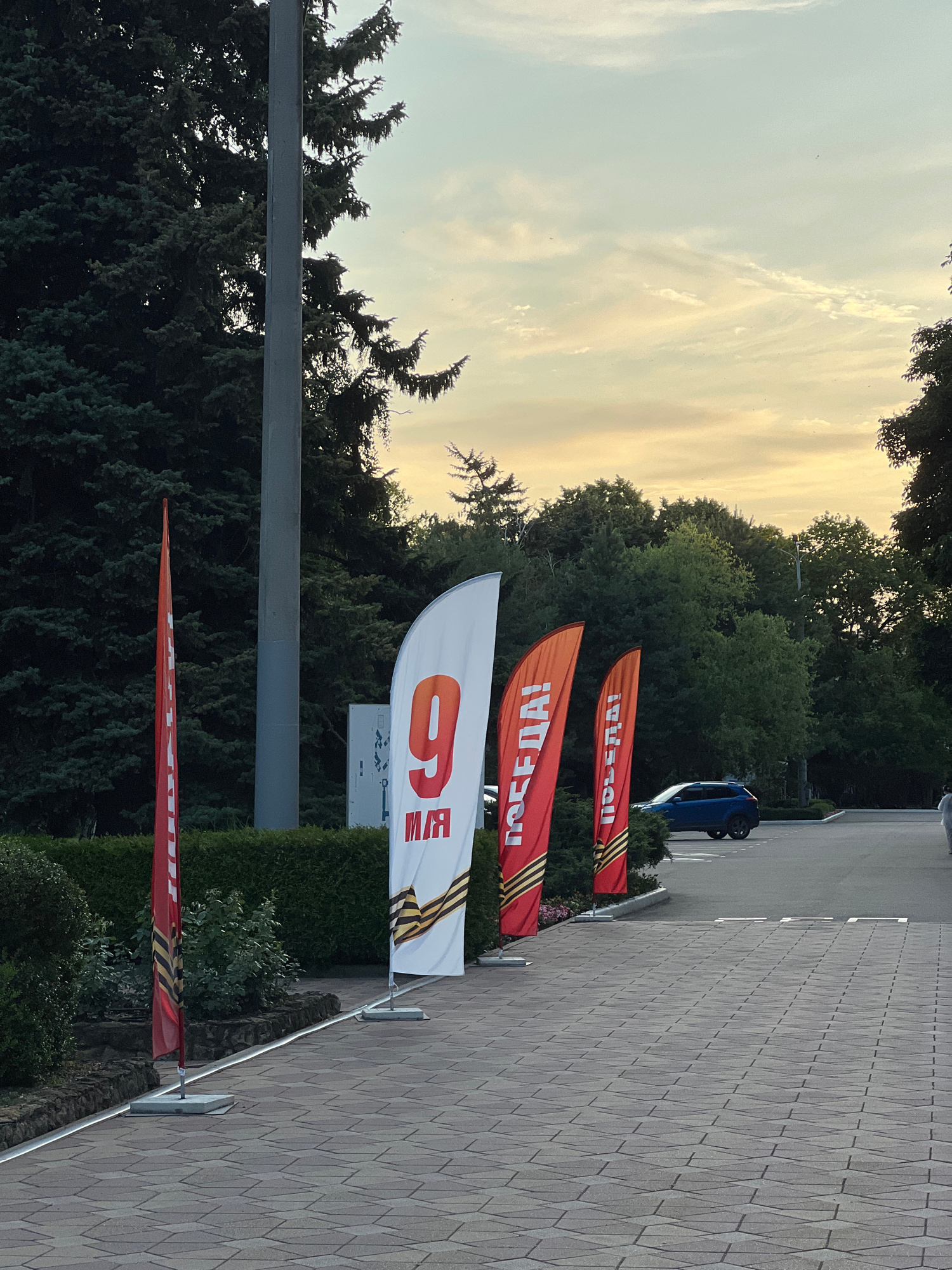 The image shows a pathway lined with several red and white flags, some featuring the number "9" and text that appears to be in Cyrillic script. One white flag has a large red "9" and text below it, along with a St. George's ribbon pattern. Two orange-red flags bear the word "ПОБЕДА!" in white Cyrillic letters, also adorned with the St. George's ribbon pattern. A thin red flag with the St. George's ribbon pattern is visible on the far left. The flags are positioned in front of a neatly trimmed green hedge. Behind the hedge, lush green trees form a backdrop, with a pale, cloudy sky visible above. A light grey utility pole stands to the left of the flags. In the background, a blue SUV is parked on an asphalt surface, and a person in light-colored clothing is walking on the right side of the frame. The pathway itself is paved with interlocking stone tiles. This scene is located in the city of Krasnodar, Russia, likely in a public space or memorial site given the commemorative flags.