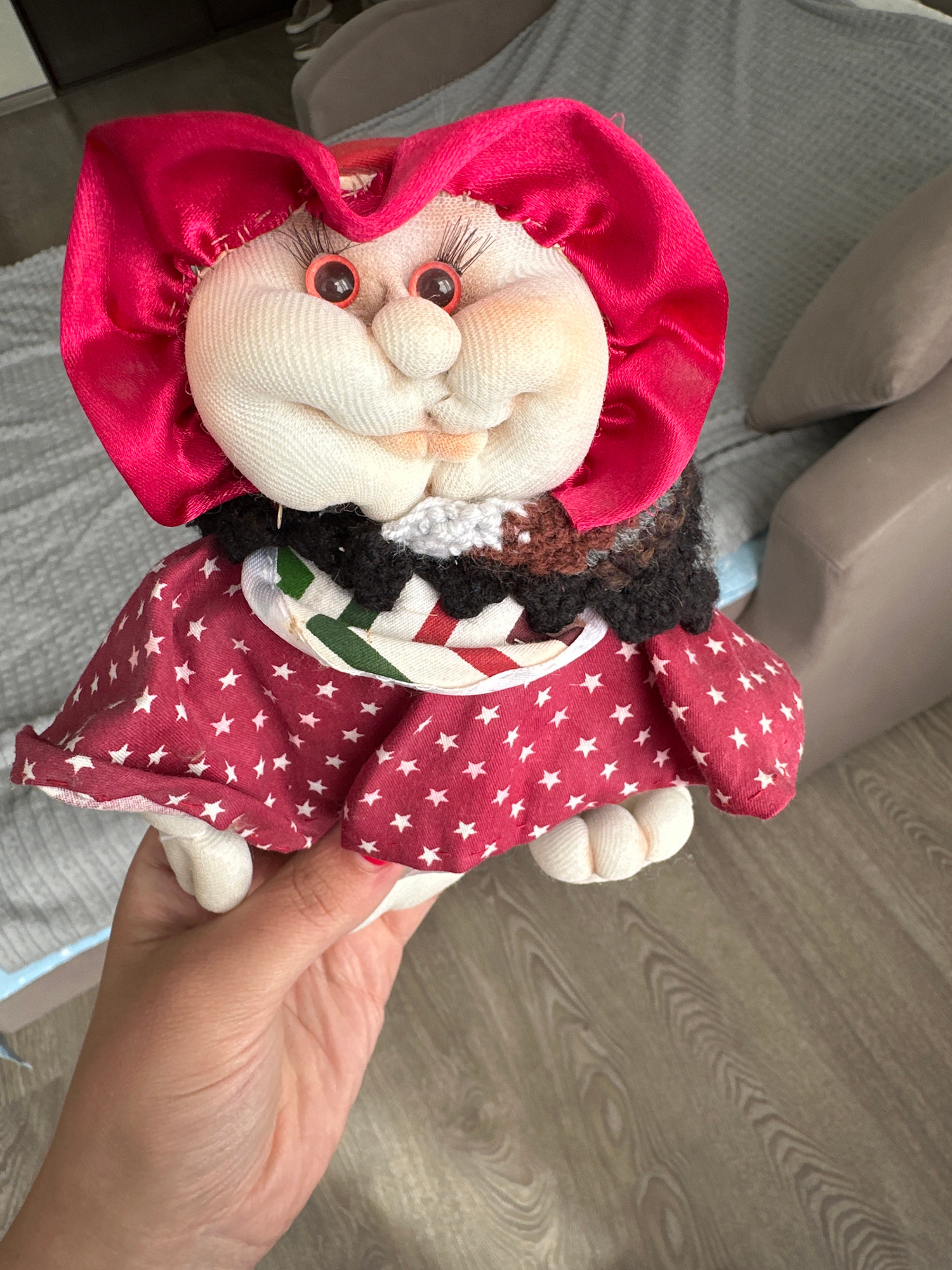 A handmade doll with a red bonnet and a red dress with white stars is held by a person's hand. The doll has round plastic eyes, a bulbous nose, and a smirking mouth. It is wearing a black and white ruffled collar. The background is blurred, showing a grey sofa and a wooden floor. The image is taken indoors, in what appears to be a residential setting. No specific location in Lytkarino, Russia, can be determined from the image.