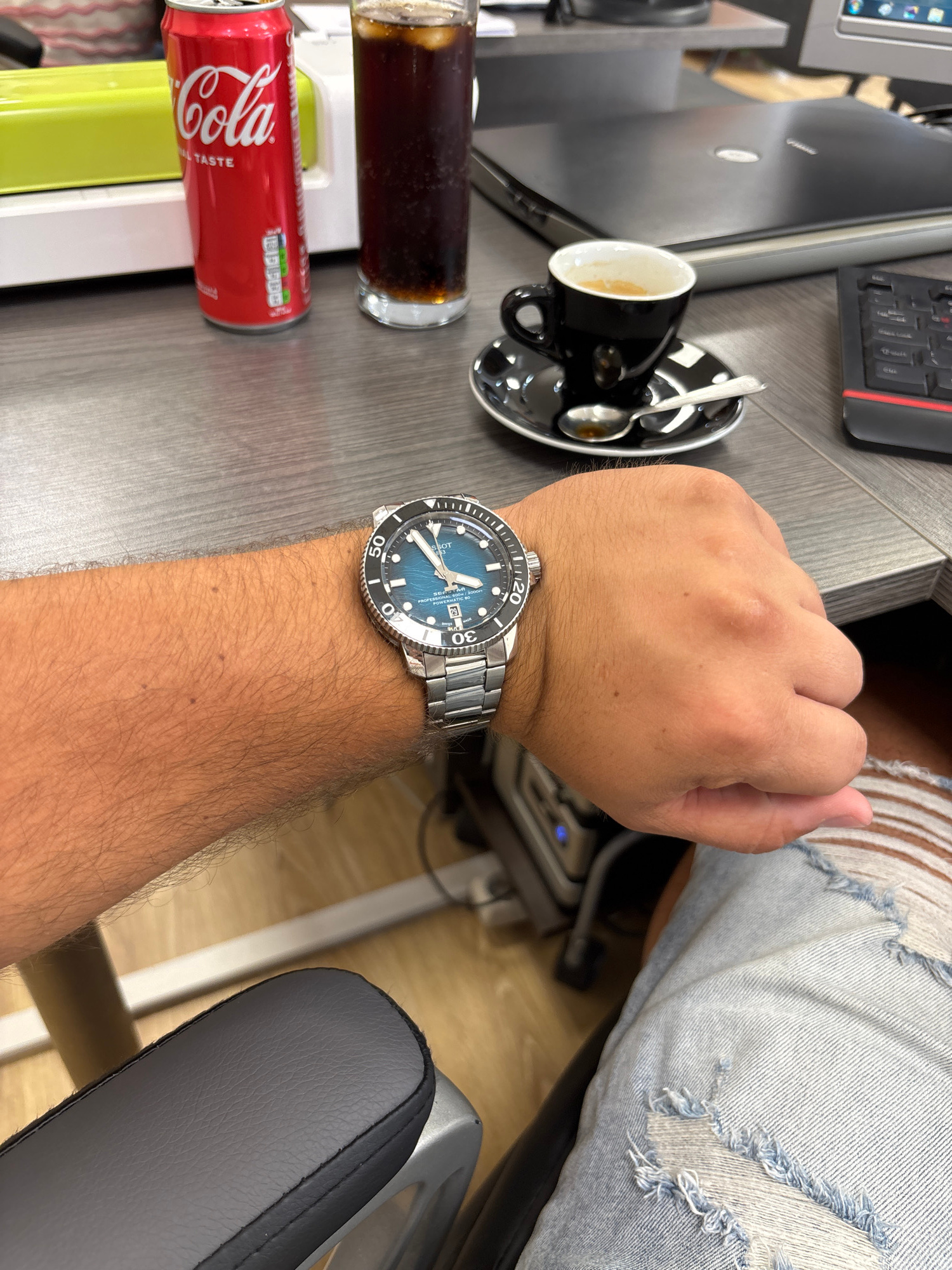 The image shows a close-up view of a person's right forearm and hand resting on a desk. The person is wearing a wristwatch with a blue dial and a metal band. On the desk, there is a red Coca-Cola can, a glass of dark liquid with ice, a black coffee cup with a saucer and a spoon, a laptop, and a keyboard. The scene appears to be an office environment. The image does not contain any information about the city of Pleven, Bulgaria.
