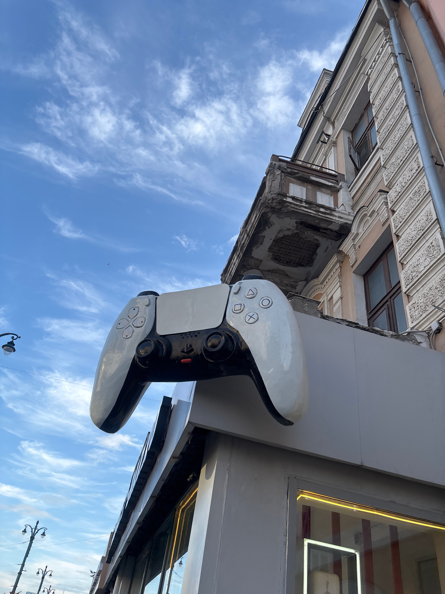 The image depicts a large, white PlayStation 5 controller mounted as a decorative element on the exterior of a building. The controller is positioned vertically, with the buttons facing the viewer. The building facade exhibits traditional architectural details, including ornate molding and weathered plaster, suggesting an older structure. Above the controller, a clear blue sky with scattered wispy clouds is visible, indicating it is daytime. The setting appears to be a street scene, with a lamppost visible in the lower right corner. The overall impression is one of urban art or decoration, likely intended to attract attention to a business or establishment associated with gaming. The image is taken from a low angle, looking upwards towards the controller and the building. There is no visible text in the image. The location context is Novaya Usman', Russia.