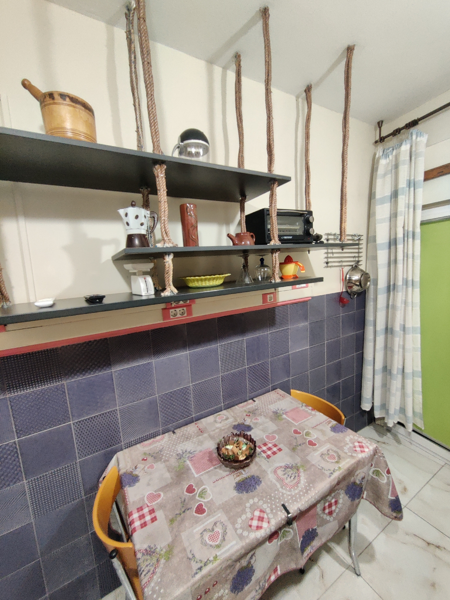 The image shows a close-up of a dining area in a room. A rectangular dining table, covered with a patterned tablecloth featuring hearts and flowers, is prominently positioned in the foreground. Two yellow chairs are placed at the table. On the table, a small bowl contains food. Behind the table, a tiled wall with blue and grey squares forms the backdrop. Above the tiled wall, a dark shelf is mounted, displaying various kitchen items including a small oven, a coffee maker, and decorative objects. Further up, another shelf is visible with a wooden bucket and a kettle. The room is lit by overhead lighting, casting shadows on the ceiling from the ropes suspending the shelves. The overall impression is of a functional and somewhat rustic kitchen or dining space. The image does not provide any explicit information to pinpoint its location within the city of Thessaloniki Municipal Unit, Greece.