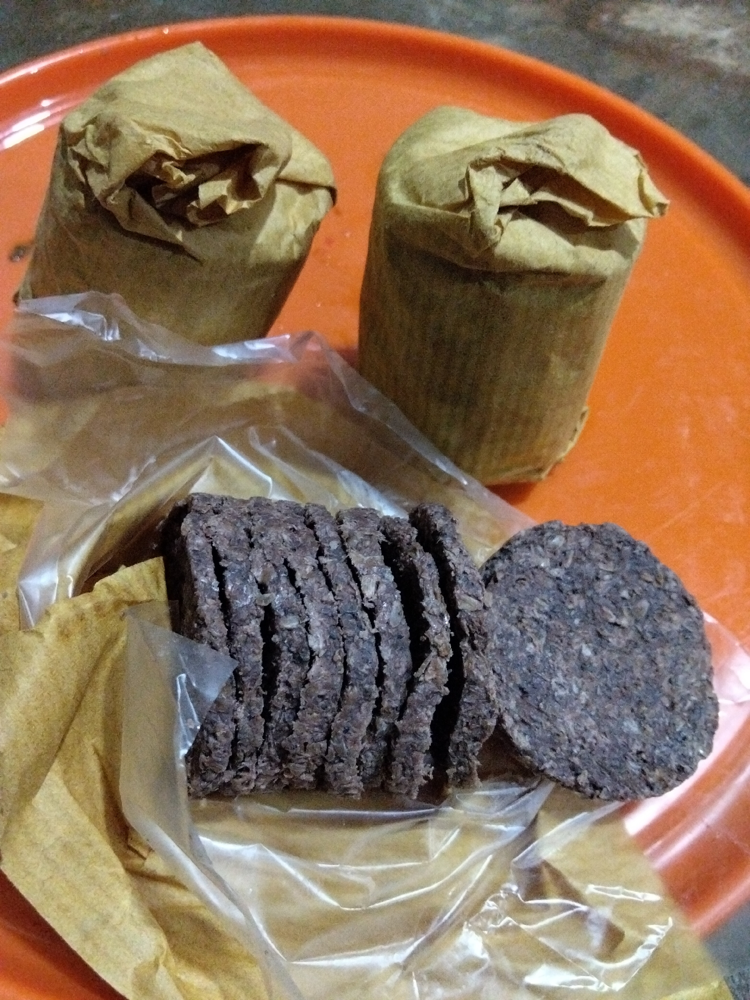 Two cylindrical objects wrapped in brown paper sit on an orange circular surface. In the foreground, a transparent plastic wrapper contains several dark brown, disc-shaped items, stacked and sliced. One disc is separate from the stack. The background is out of focus.