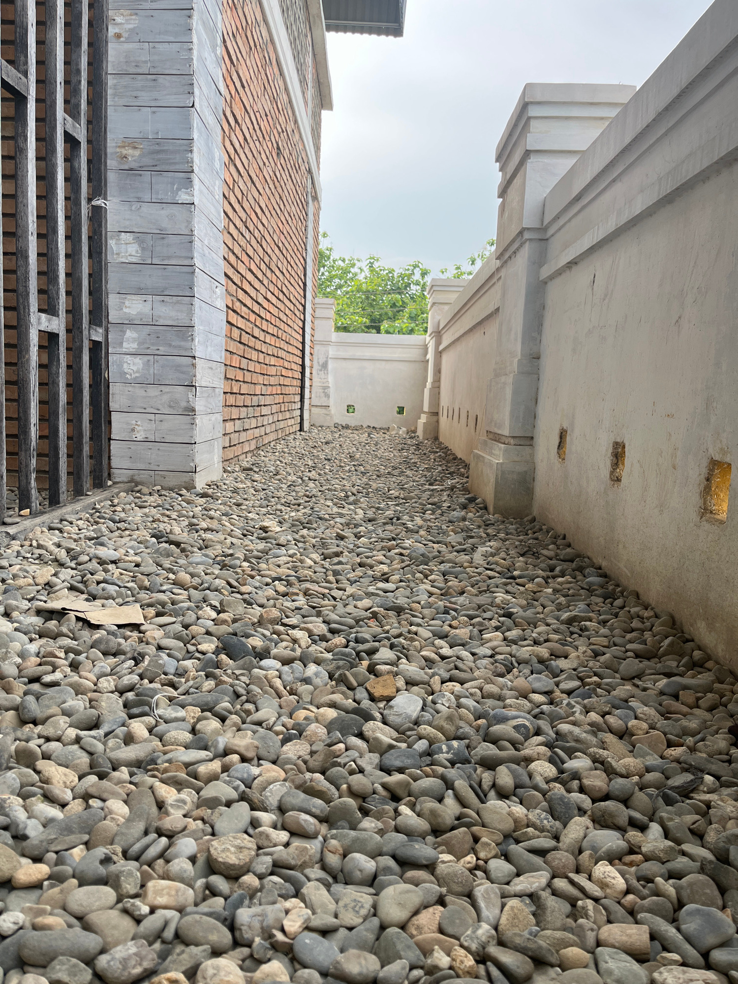 The image depicts a narrow pathway lined with pebble-like stones, leading between two structures. On the left, a brick wall is visible, partially covered with weathered wooden planks and adjacent to a dark metal fence. On the right, a smooth, light-colored stucco wall with decorative columnar elements and recessed square openings runs alongside the path. The sky above is overcast. The scene is devoid of any people or animals. The location is identified as being in Benuaraja, Indonesia.