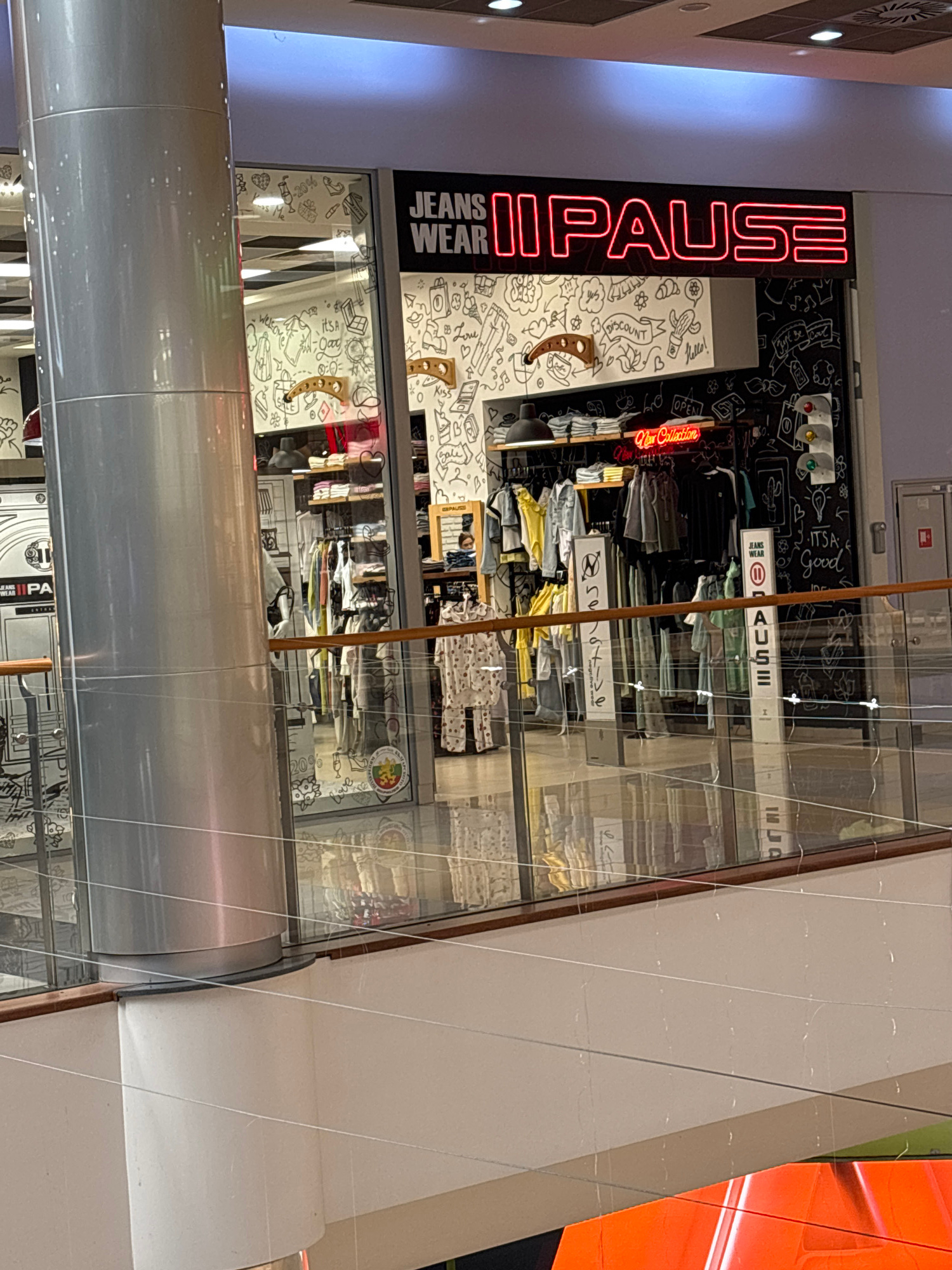 The image shows the storefront of a clothing store called "IPAUSE," which specializes in jeans and casual wear. The store features a prominent neon sign spelling out "IPAUSE" in red letters vertically along the left side of the entrance. The storefront has large glass windows displaying racks of clothing, including t-shirts, jeans, and other apparel. Some of the clothing is neatly folded, while other items are hung on racks.
The interior of the store is decorated with black and white doodle-style artwork on the walls, featuring various images and text such as "IT'S Good," "LOVE," and "Kiss." There are also signs within the store, including one that says "OPEN" and another that says "Car Collection." Small tags and price information are visible on some of the clothing items.
The overall scene suggests a retail environment, likely a shopping mall or a street-level store in an urban setting. The lighting indicates it could be daytime or evening, with artificial lights illuminating the store's interior and exterior signage. The image is captured from a slightly upward angle, showing the facade of the store and a portion of the surrounding structure, which appears to be part of a modern building with metallic elements. There are no people visible in the frame, but the presence of merchandise suggests the store is open for business. The location context provided is Pleven, Bulgaria.