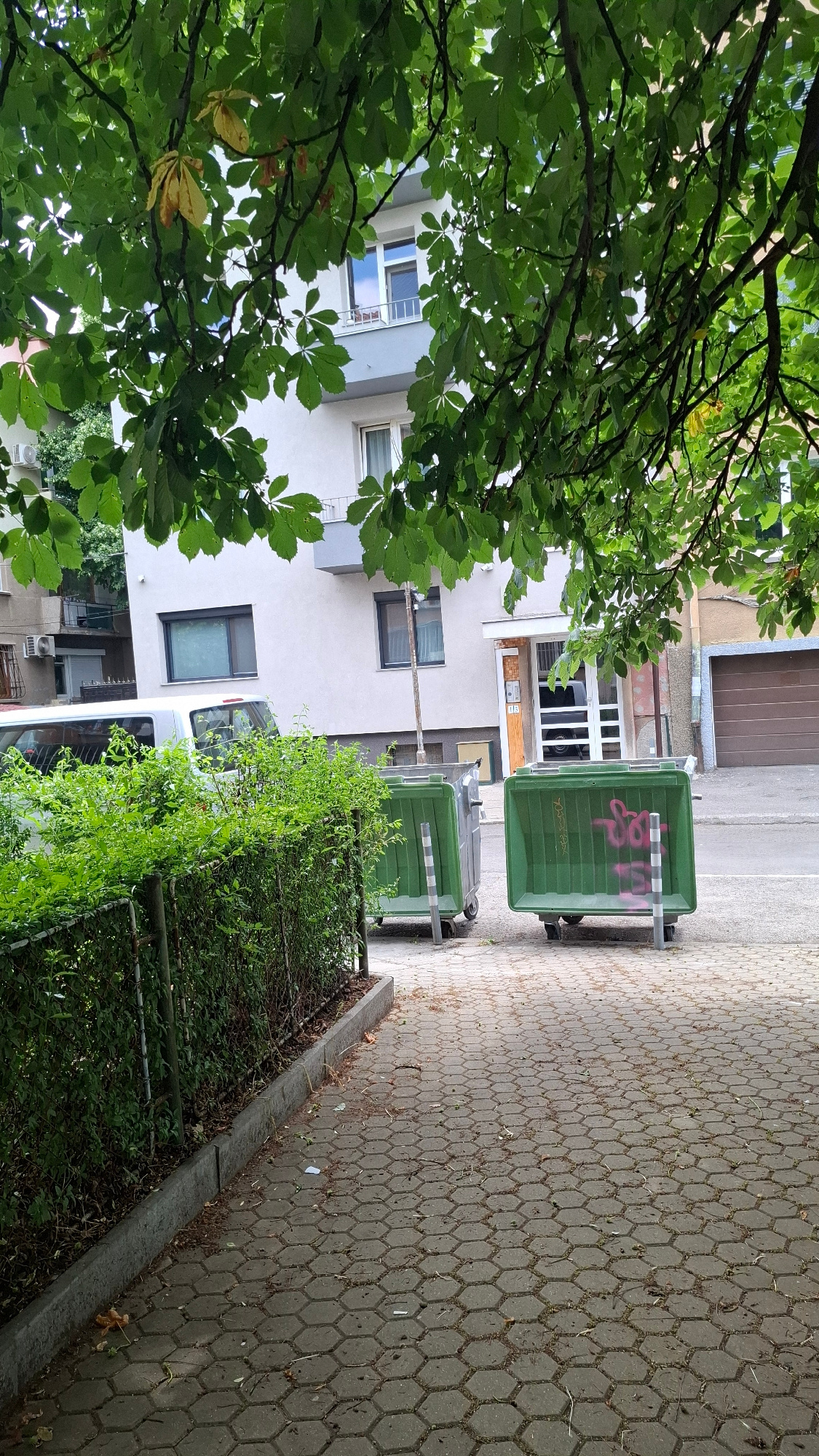 In Sofia, Bulgaria, on a paved sidewalk with a hexagonal pattern, two green dumpsters stand side-by-side. The dumpster on the right is marked with pink graffiti. Behind the dumpsters, a grey building with multiple windows and balconies is visible. A silver van is parked to the left of the dumpsters, partially obscured by a dense green hedge that runs along a fence. Overhead, the branches of a tree with green and yellowing leaves cast shadows. The scene appears to be an outdoor urban setting.