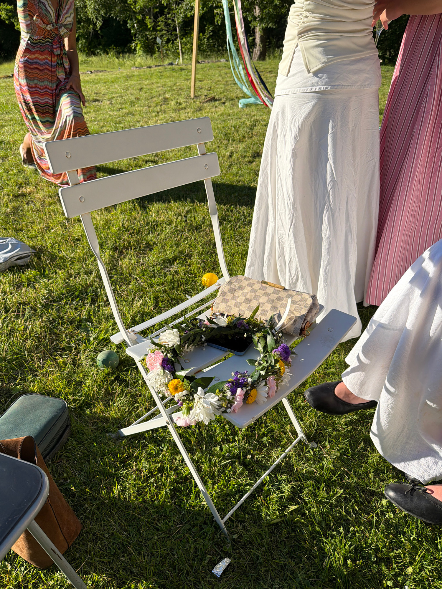 A close-up, high-angle shot captures a white folding chair situated on a sunlit grassy lawn. The chair is adorned with a floral arrangement, a smartphone, and a designer clutch purse. In the background, three individuals are partially visible, wearing light-colored dresses and skirts, with one person in a pink and white striped skirt. The setting appears to be an outdoor gathering, possibly a garden party or event. The image does not contain specific landmarks or indicators to pinpoint a location within Stockholm, Sweden.