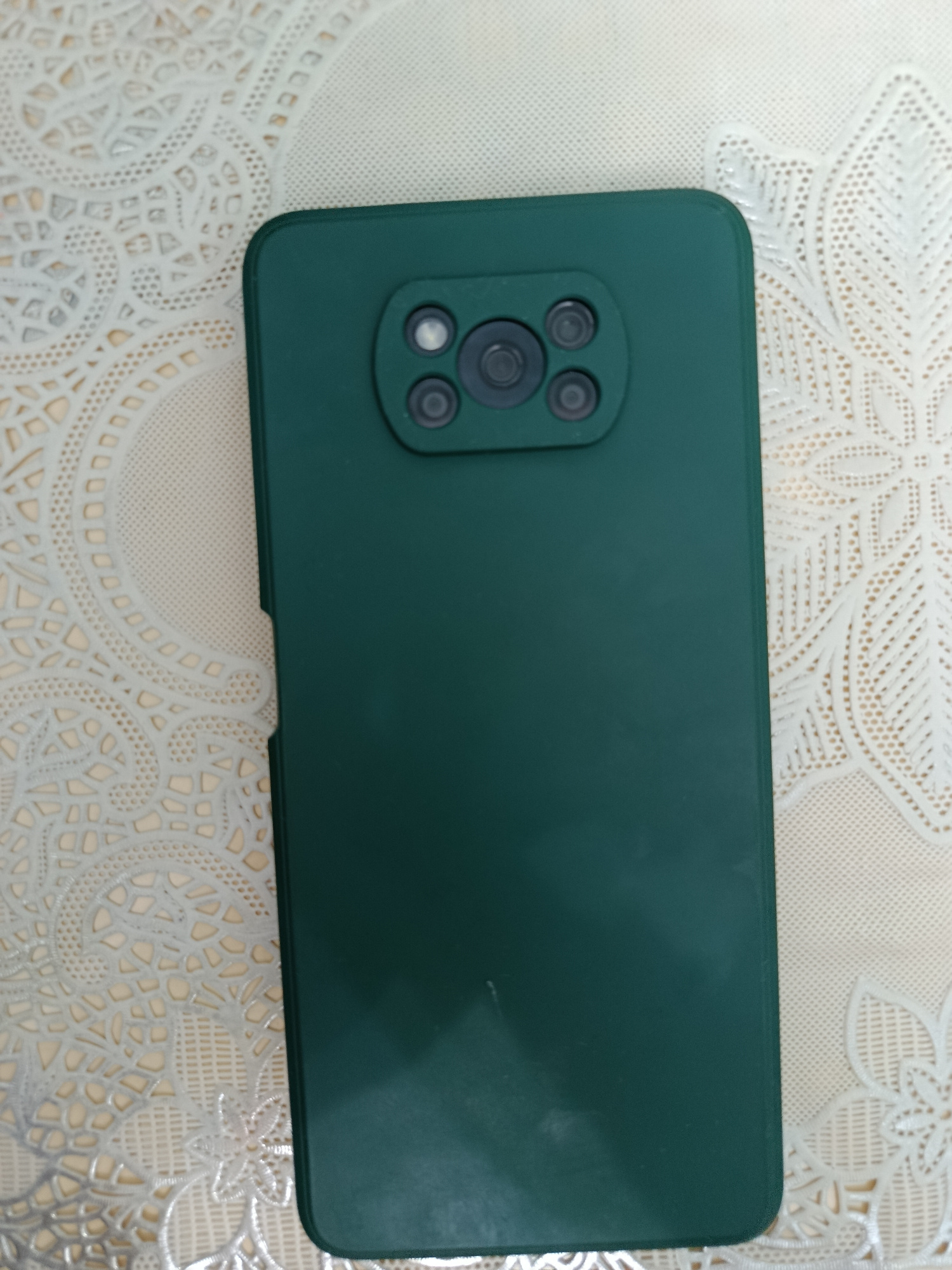 A dark green smartphone with a camera array is placed on a cream-colored tablecloth with a white floral pattern. The smartphone is positioned vertically, with the camera module at the top. The camera module consists of four circular lenses arranged in a square formation, with a flash element visible in the upper right portion of the module. The smartphone is partially covering the tablecloth. The lighting is even, and there are no visible shadows indicating a specific light source. The background is a plain cream color, with the textured pattern of the tablecloth providing visual interest. The image is taken from a slightly overhead angle, looking down at the smartphone and tablecloth. There are no people or any other objects visible in the image. The scene depicts a still life composition of a single electronic device on a decorative fabric. It's not possible to determine the location in Tashkent, Uzbekistan from the provided image.