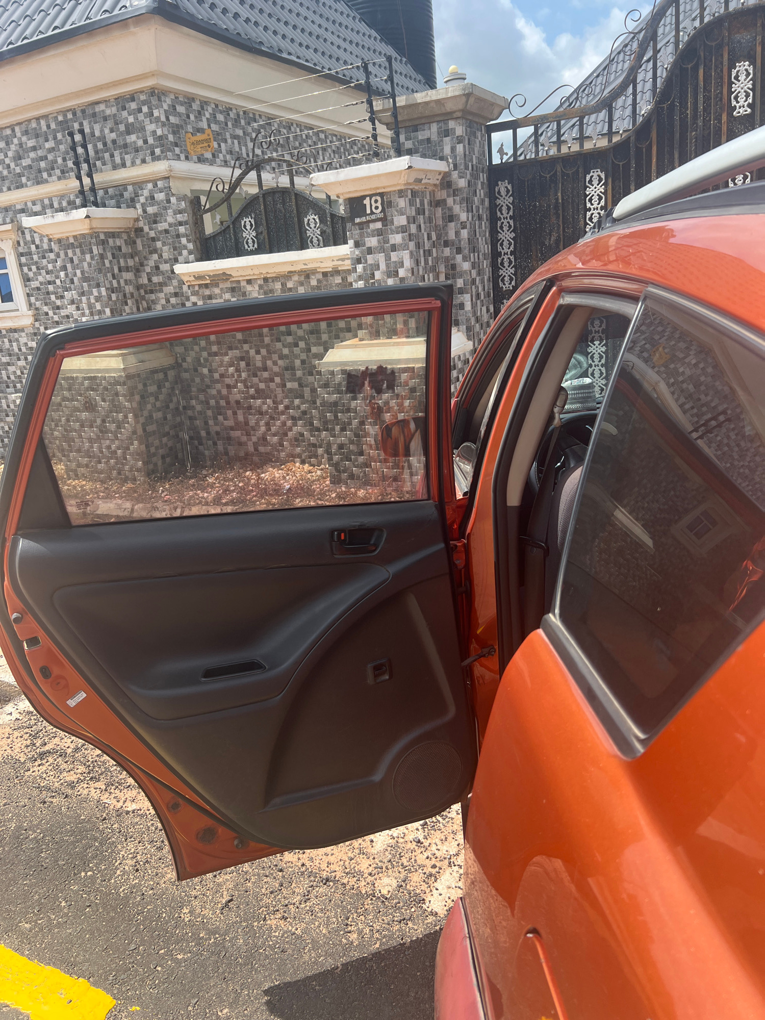 An orange car door is open, revealing the interior of the vehicle and the surrounding environment. The car is parked on an asphalt surface with a yellow line marking. Behind the car, a house with a tiled facade is visible. The house has a fence with decorative metalwork and a sign that reads "18". The sky is cloudy. The image appears to have been taken during the daytime. The scene is static, with no people or action visible. The focus is on the car door and the immediate surroundings.