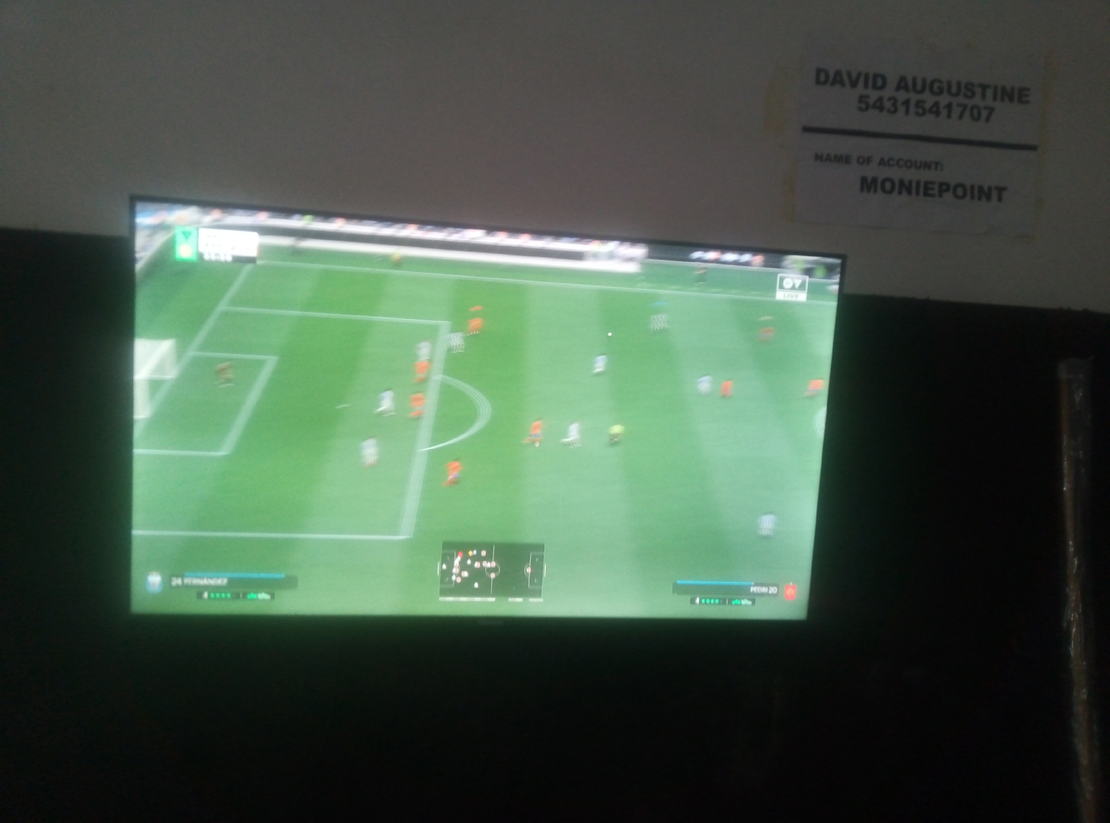 The image captures a television screen displaying a soccer video game, likely FIFA or a similar simulation. The game features a top-down view of a green soccer field with numerous players in various colored kits. A mini-map of the field is visible in the lower left portion of the screen. In the upper right corner of the image, a paper is affixed to a wall, with text printed in black ink. The text includes a name, "DAVID AUGUSTINE," followed by a series of numbers, "5431541707." Below this, it reads, "NAME OF ACCOUNT:" and then "MONIEPOINT." The scene appears to be indoors, with the television screen prominently positioned in the foreground. The lighting suggests a dimly lit environment, possibly a room or an establishment. The wall texture is visible behind the paper. The overall impression is that of a sports viewing area where financial transaction information is displayed alongside a game.