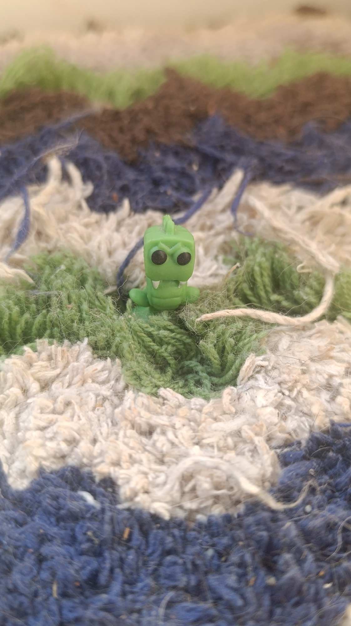 A small, bright green toy dinosaur figurine sits on a textured rug with horizontal stripes of color. The dinosaur is cartoonish, with large, round black eyes and a wide, open mouth showing small white teeth. It has a crest of three points on its head and two stubby arms. The rug has alternating wavy bands of green, brown, and dark blue, with some cream-colored fibers interspersed. The lighting is bright, suggesting it might be daytime, but the exact time of day and location are unknown. The shallow depth of field blurs the background and the rug fibers surrounding the figurine, drawing attention to the toy itself. No text is visible in the image.