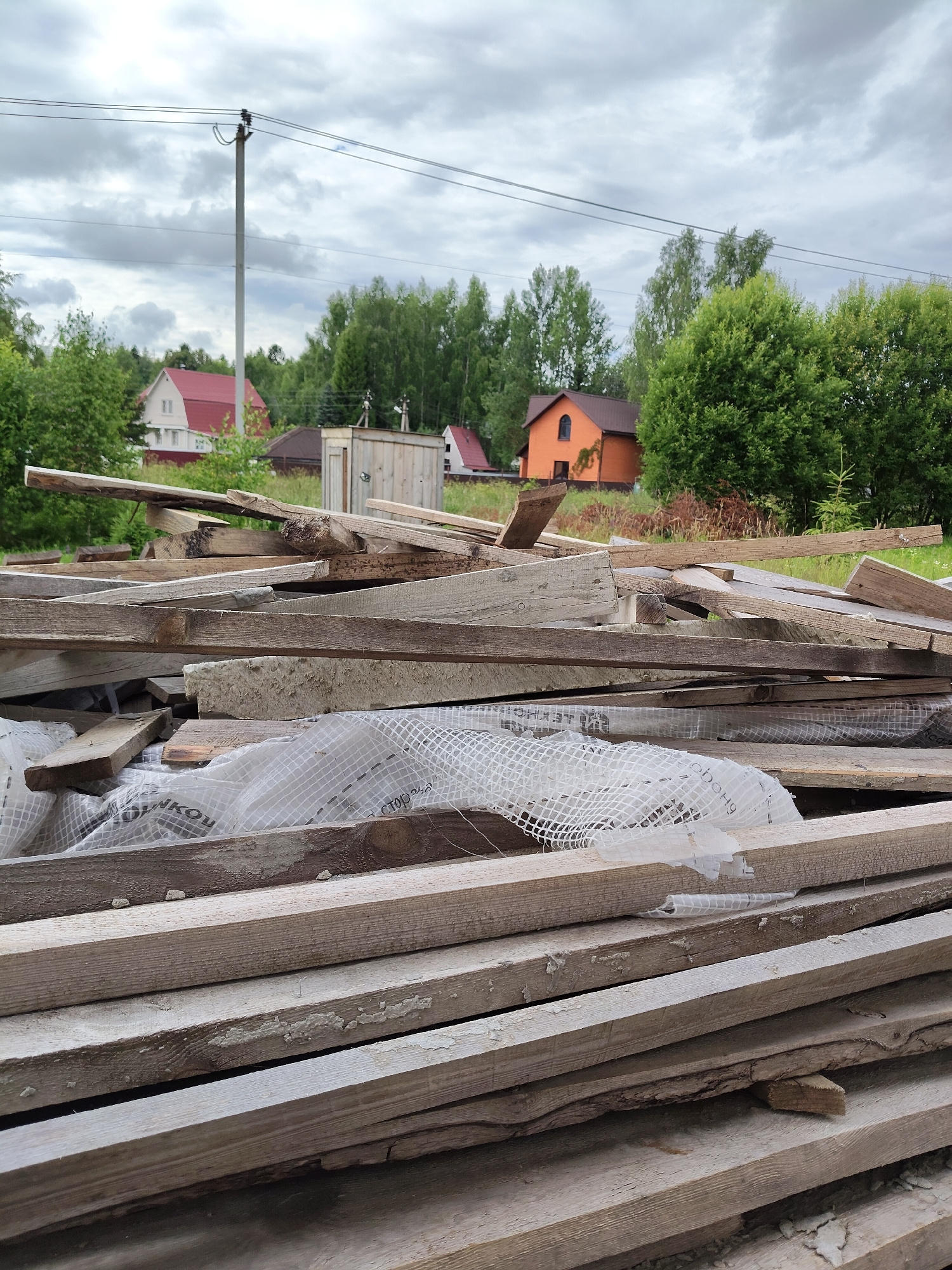 This media file depicts a pile of discarded wooden planks in an outdoor setting, likely a construction or renovation site. The planks are of various sizes and appear weathered, with some showing signs of decay and peeling paint. A white, mesh-like material is partially visible underneath some of the planks, possibly a protective covering.
In the background, several houses are visible, suggesting a semi-rural or suburban location. One house has a prominent red roof, while another is painted a bright orange. Lush green trees and bushes surround the structures, indicating a season with active foliage, likely summer or late spring. A utility pole with overhead wires stands tall in the middle ground, further contextualizing the environment.
The sky is overcast with heavy, grey clouds, suggesting a cloudy or impending rainy day. The overall lighting is diffused, and there are no strong shadows. The scene appears still, with no visible people or animals. The focus is primarily on the pile of wood in the foreground, emphasizing its disarray.
Some text is visible on the white mesh material. Though partially obscured and blurry, it appears to be in Cyrillic script. Phrases like "СТОРОНА" (meaning "side") and other partial words like "ЯКО" can be discerned. The location context provided is Скурыгино, Russia.