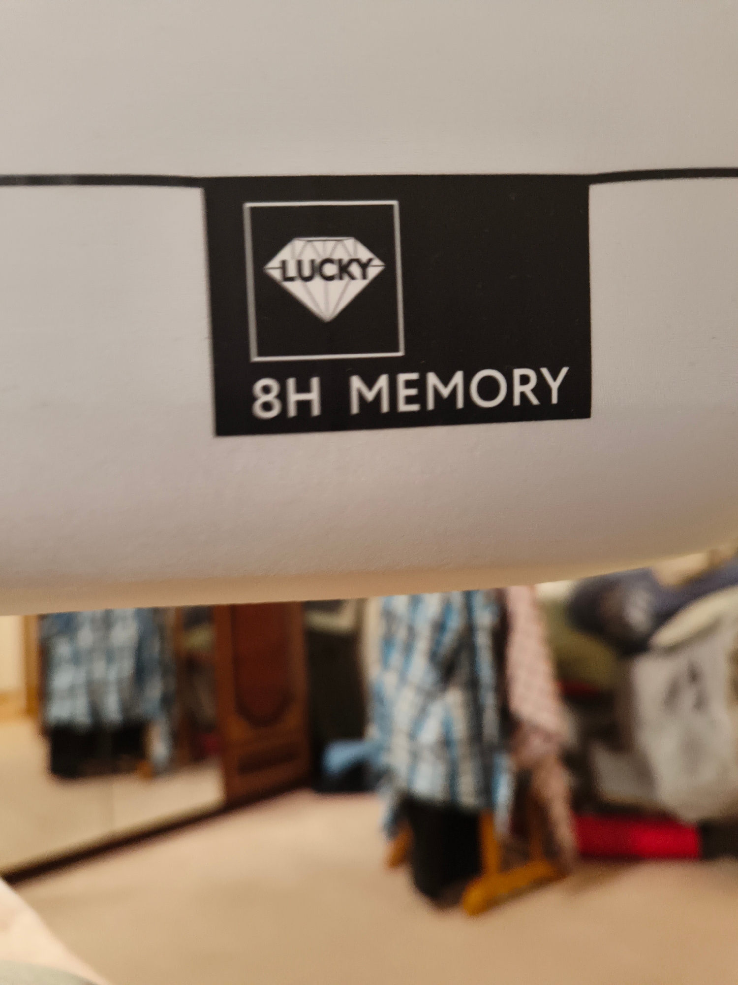 The image displays a close-up of a white surface with a black rectangular label. The label features a white outline of a diamond with the word "LUCKY" written above it in white text. Below the diamond graphic, the text "8H MEMORY" is written in white, capital letters. The background is blurred and appears to be an indoor environment with furniture and clothing visible. There are no people present in the immediate frame of the label. The overall scene suggests a product label or branding element. There is no information to indicate the location in the city of Seversk, Russia.