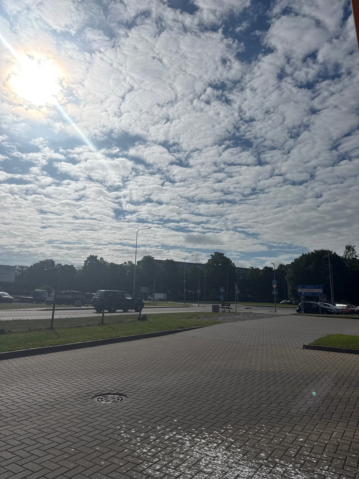 The image captures a scene from ground level, oriented vertically. The sky dominates the left side, filled with a vast expanse of clouds. These clouds are light and fluffy, arranged in horizontal bands across the blue sky, suggesting a partially cloudy day. The sun is visible on the far left, casting a bright glare and a sunbeam that cuts diagonally across the sky and clouds.
The right side of the image shows a paved walkway made of dark gray bricks arranged in a herringbone pattern. There are some white splotches on the bricks, possibly from water or debris. Adjacent to the walkway is a strip of green grass, and beyond that, a chain-link fence.
Behind the fence, there are trees with dark green foliage. Several cars are parked in a lot or along a road next to the trees. A street light is also visible, as is a road sign with directions indicated by arrows. Some text is visible on the road sign, appearing to be in a non-English language, with words like "RAMUČIAI 10" and "CENTRAS". There is also a round metal grate set into the pavement of the walkway. The overall impression is a bright, somewhat overcast day in an urban or suburban setting, possibly during the daytime due to the sunlight. There are no people clearly visible in the frame.