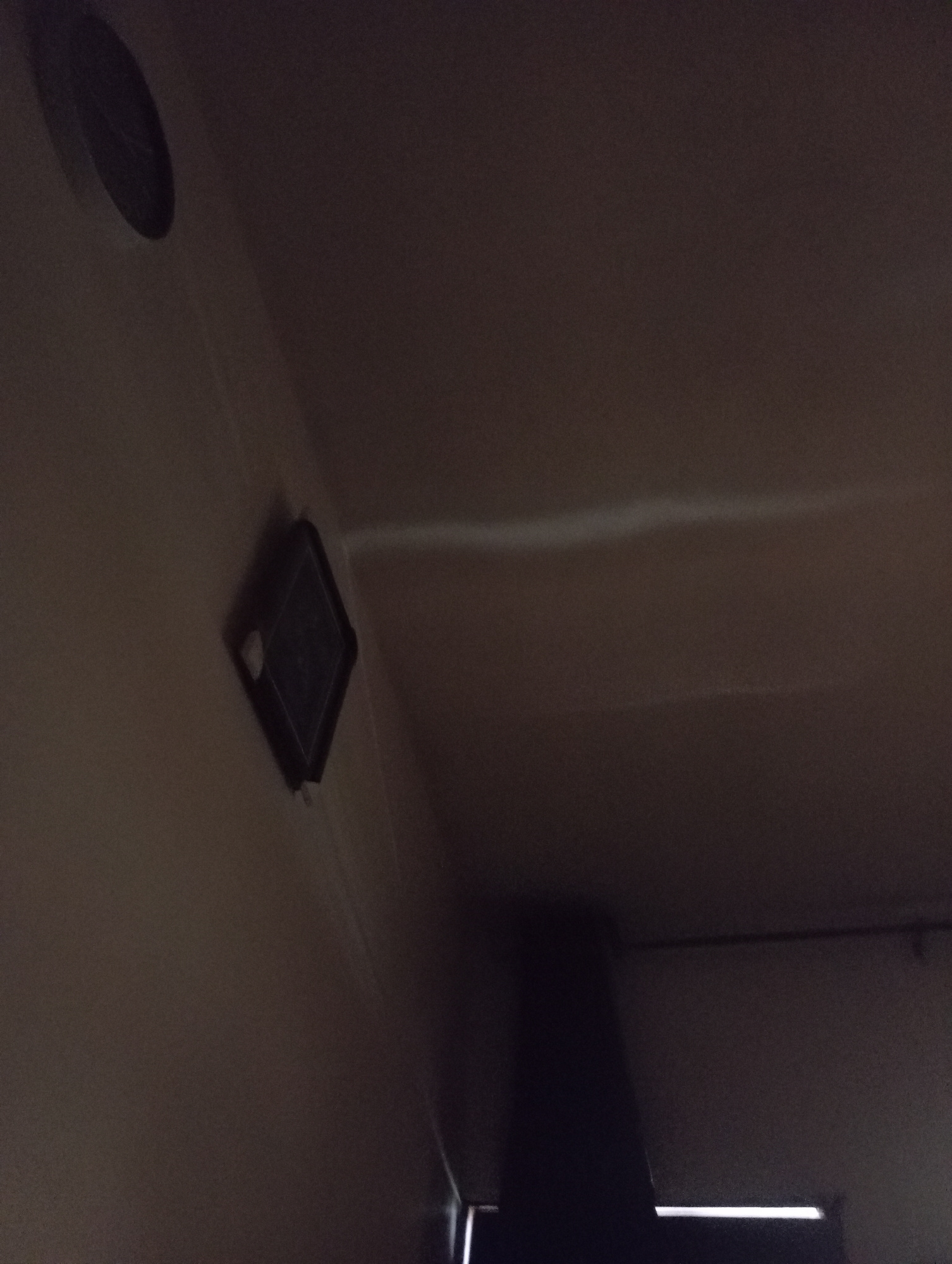 A dark, dimly lit indoor scene captures a ceiling-mounted diamond-shaped object, possibly a detector or sensor, with a faint white light emanating from its center. To the left of this object, a circular ventilation grate is visible, also mounted on the ceiling. The walls and ceiling are a neutral, muted color, suggesting a residential or commercial interior. The lighting is low, creating shadows and obscuring finer details. The overall impression is one of an ordinary interior space, possibly a hallway or room, in Nkpor, Nigeria.