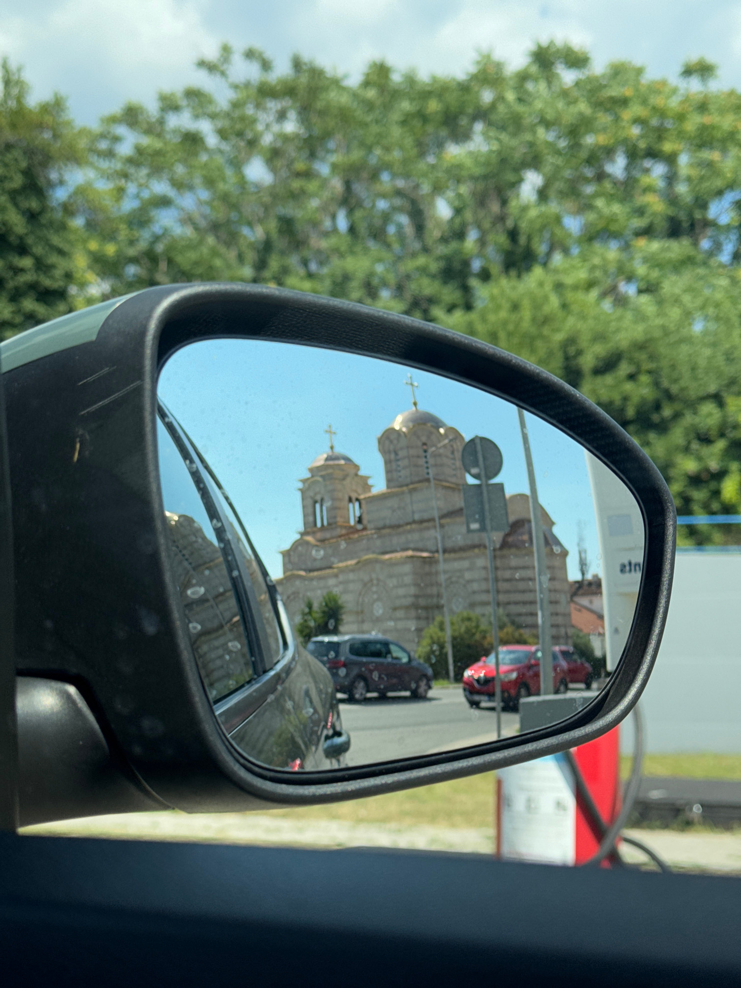 This media file shows a reflection in a car's side mirror. The reflection features a large, ornate building with multiple domes, characteristic of a church or cathedral, with golden crosses atop the domes. The building appears to be made of stone and has intricate architectural details.
In front of the building, a street is visible, with several cars parked or driving. One red car is prominently featured in the reflection. The sky in the reflection is clear and blue, suggesting daytime.
The mirror itself is framed in black, with a textured surface that appears to be carbon fiber. Outside the mirror, in the foreground and background of the actual image, lush green trees and foliage are visible, indicating an outdoor setting with ample greenery. The overall scene suggests a sunny day.
The setting is likely in Tivat, Montenegro, as indicated by the context. The presence of a church with domes points to a location with a rich religious or historical architectural heritage. The reflection in the mirror captures a moment in time, showing a street scene with vehicles and a significant architectural landmark. There are no people directly visible in the reflection. Visible text in the reflection appears to be a brand name on a sign in the background, "eam" is visible.