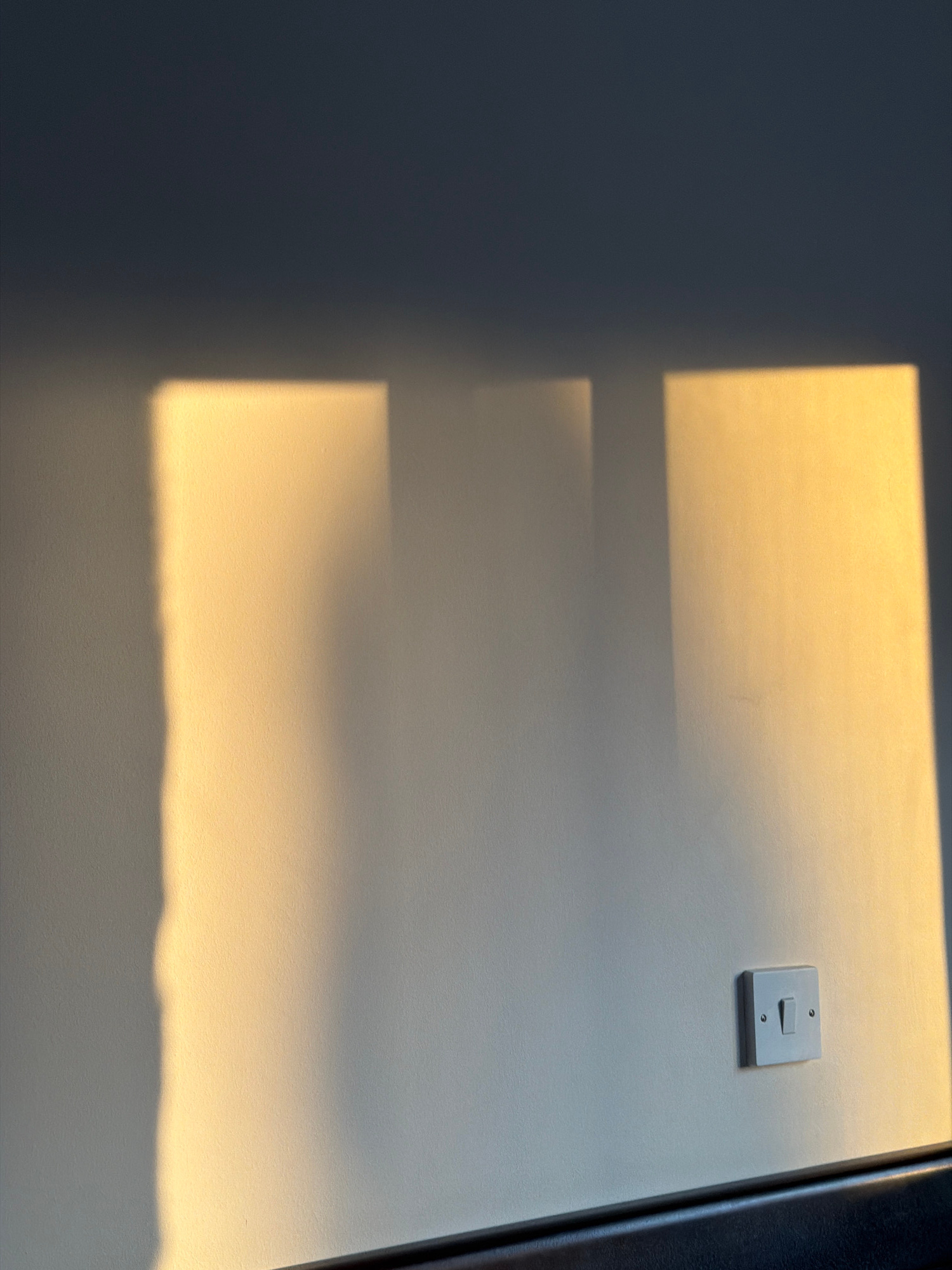 The image depicts a plain, light-colored wall with distinct geometric shadows cast upon it, indicative of sunlight entering through a window or blinds. The shadows are a warm yellow-orange hue, suggesting the golden hour of either sunrise or sunset. The wall has a slightly textured surface. In the lower right quadrant of the image, a standard white light switch is mounted on the wall, secured by two screws. The switch is in the "off" position. A dark, horizontal surface, possibly a windowsill or furniture, runs along the bottom edge of the frame. The image does not contain any people or animals. The environment appears to be indoors, in a room with a wall, light switch, and a window. The location in Tersefanou, Cyprus, is not visually discernible from the image itself.