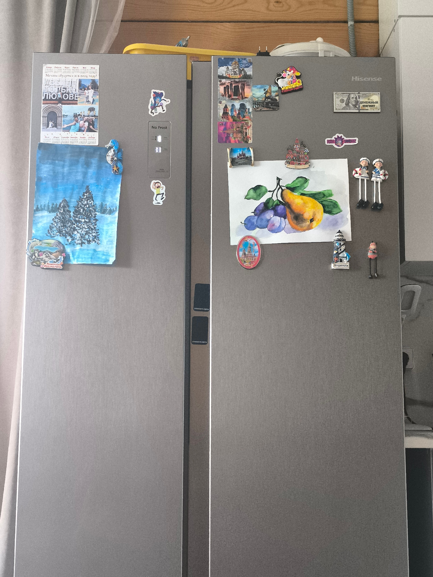 The image shows a Hisense refrigerator in a room with light-colored walls and a wooden ceiling. The refrigerator doors are decorated with various magnets and a painting. The left door has a painting of two snow-covered trees in a winter landscape, a magnet with a seahorse, and a collage of photos and text. The right door features a watercolor painting of a pear and plums, a magnet shaped like a lighthouse, a small figurine of a woman, a magnet with two sailor figures, a magnet depicting a building with a green dome, and a magnet in the shape of a dollar bill with Russian text. There are also several other smaller magnets on this door, including one with a cartoon character. The refrigerator is situated in a domestic setting, likely a kitchen. There is no specific indication of the city of Khomutovo, Russia, in the visual details provided.