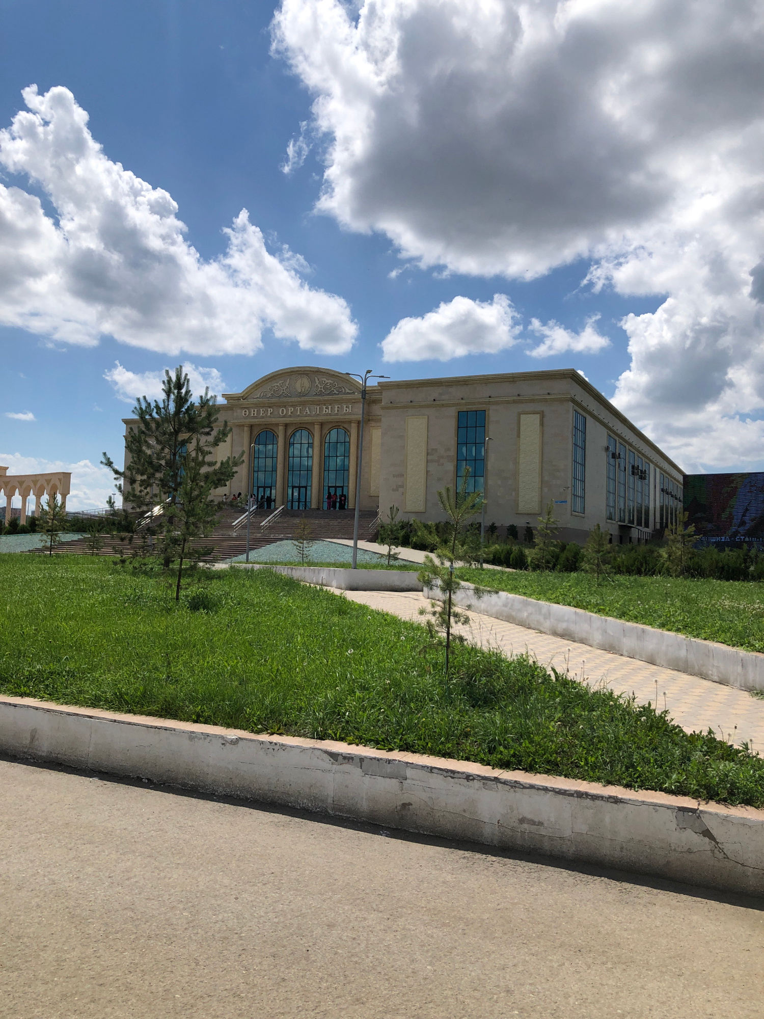 A contemporary building with a light-colored facade and tall, arched windows stands under a partly cloudy sky. The building appears to be a cultural center or museum, with the words "ӨНЕР ОРТАЛЫҒЫ" inscribed above the entrance. The foreground features a well-maintained lawn and a paved walkway. In the distance, an arched colonnade is visible, adding to the architectural interest of the scene. The overall impression is one of a modern, well-kept public space.